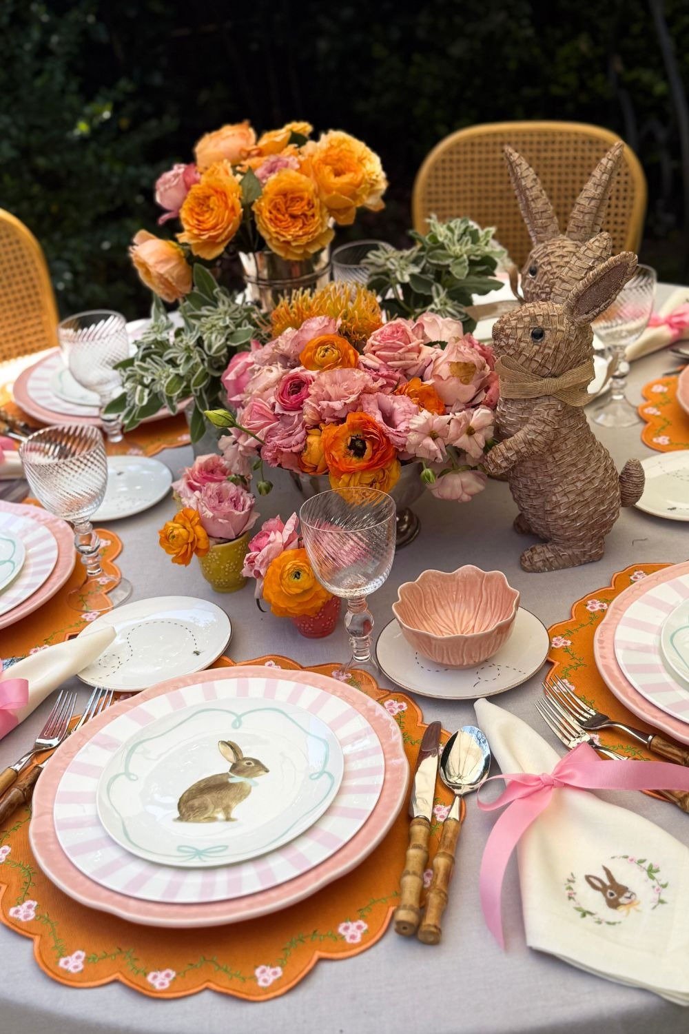 Mesa posta para Páscoa com louça rosa de coelho, flores vibrantes, sousplats laranja e clima acolhedor ao ar livre