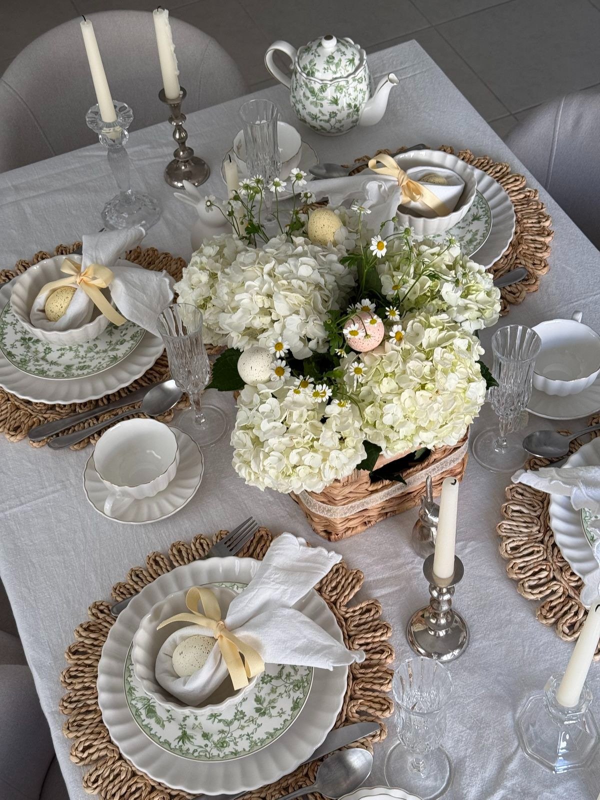 Mesa posta para Páscoa com louça branca e verde, sousplats de palha, flores brancas, velas e laços amarelos delicados