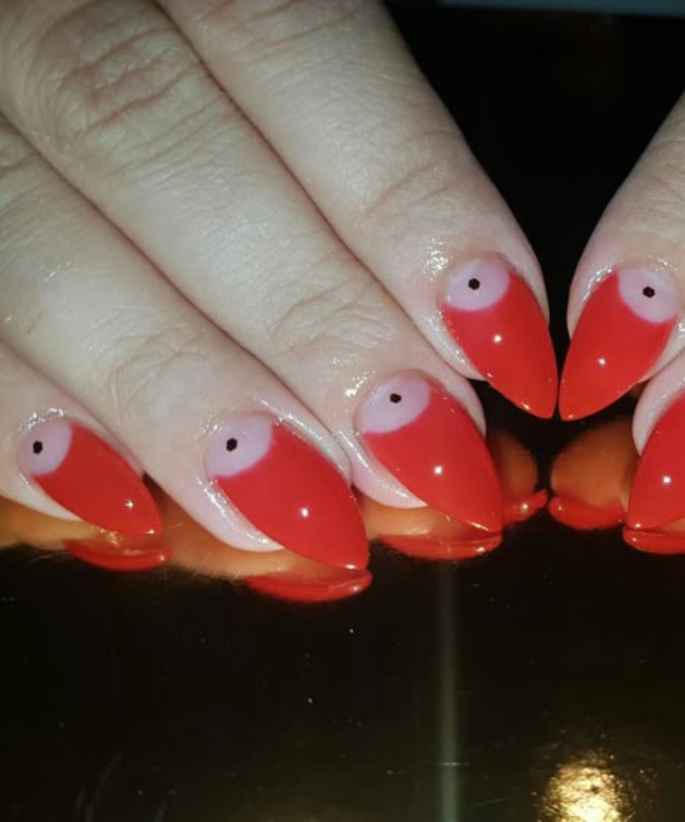Esmaltação polêmica com unhas pontiagudas em vermelho vibrante e meia-lua rosa claro com ponto preto, criando contraste