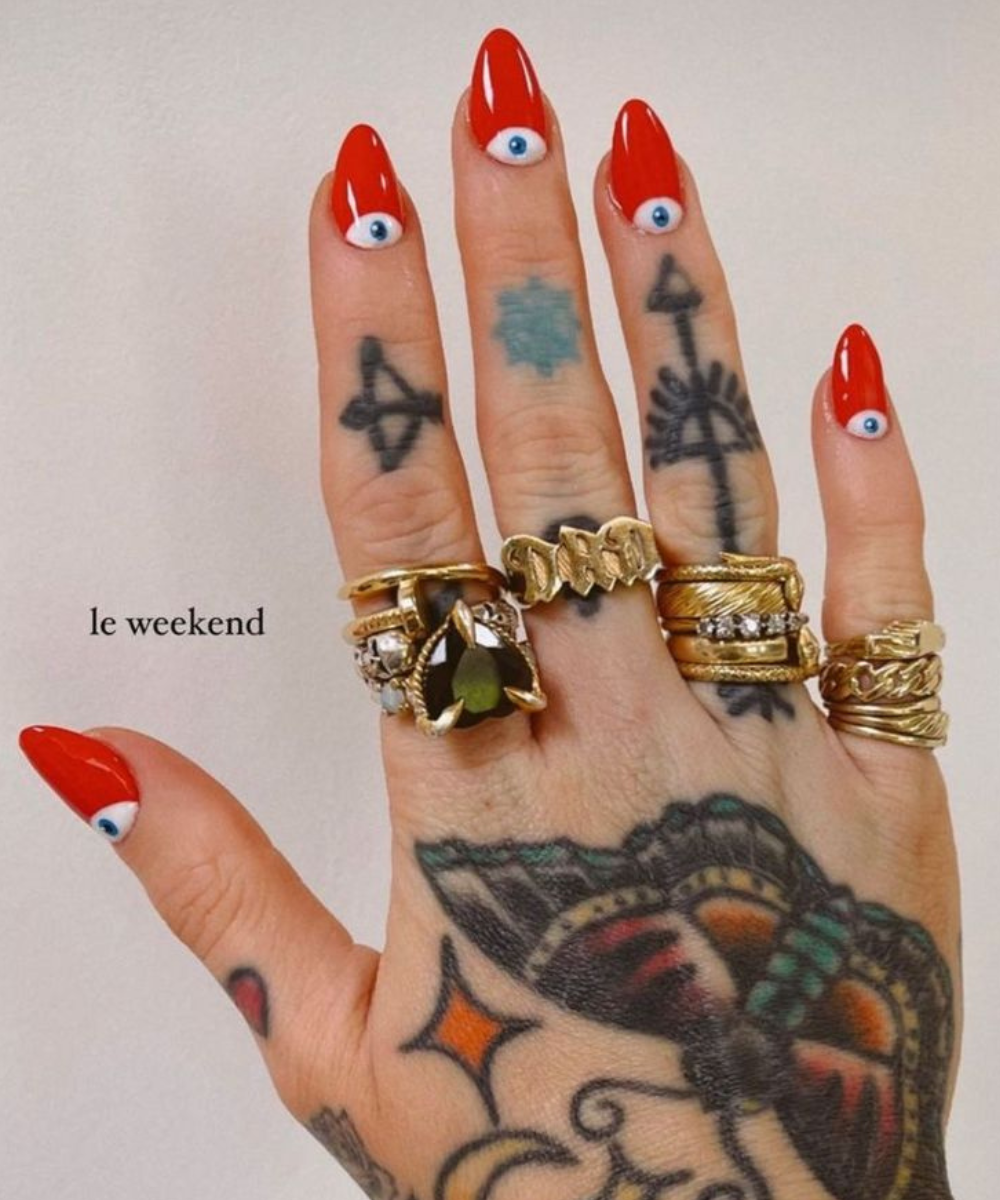 Mão tatuada exibe esmaltação polêmica em vermelho vibrante com olhos 3D, adornada por anéis dourados robustos e design