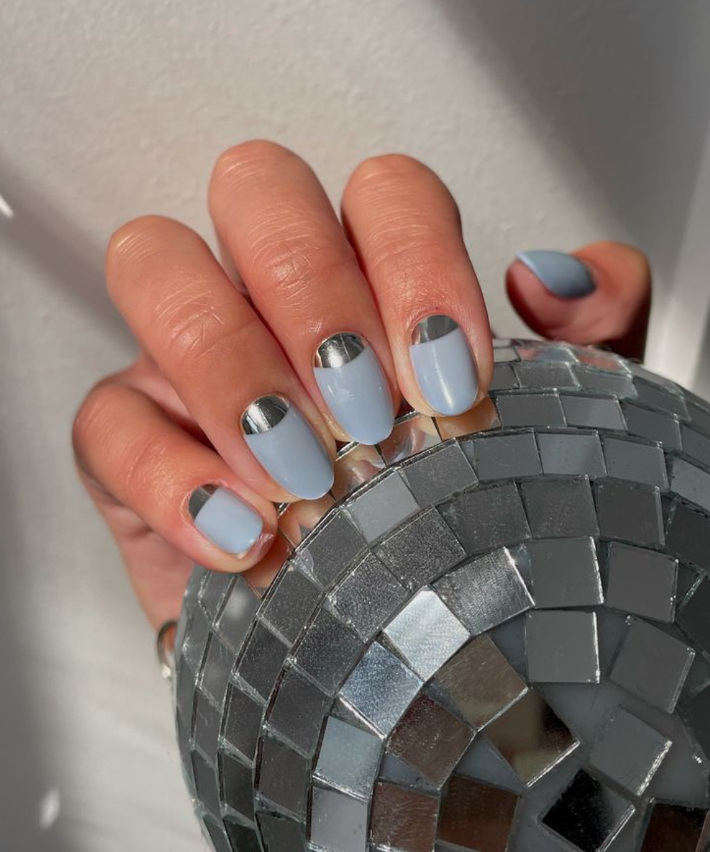 Esmaltação polêmica com base azul clara e meia-lua espelhada, unindo modernidade e brilho em unhas ovais.