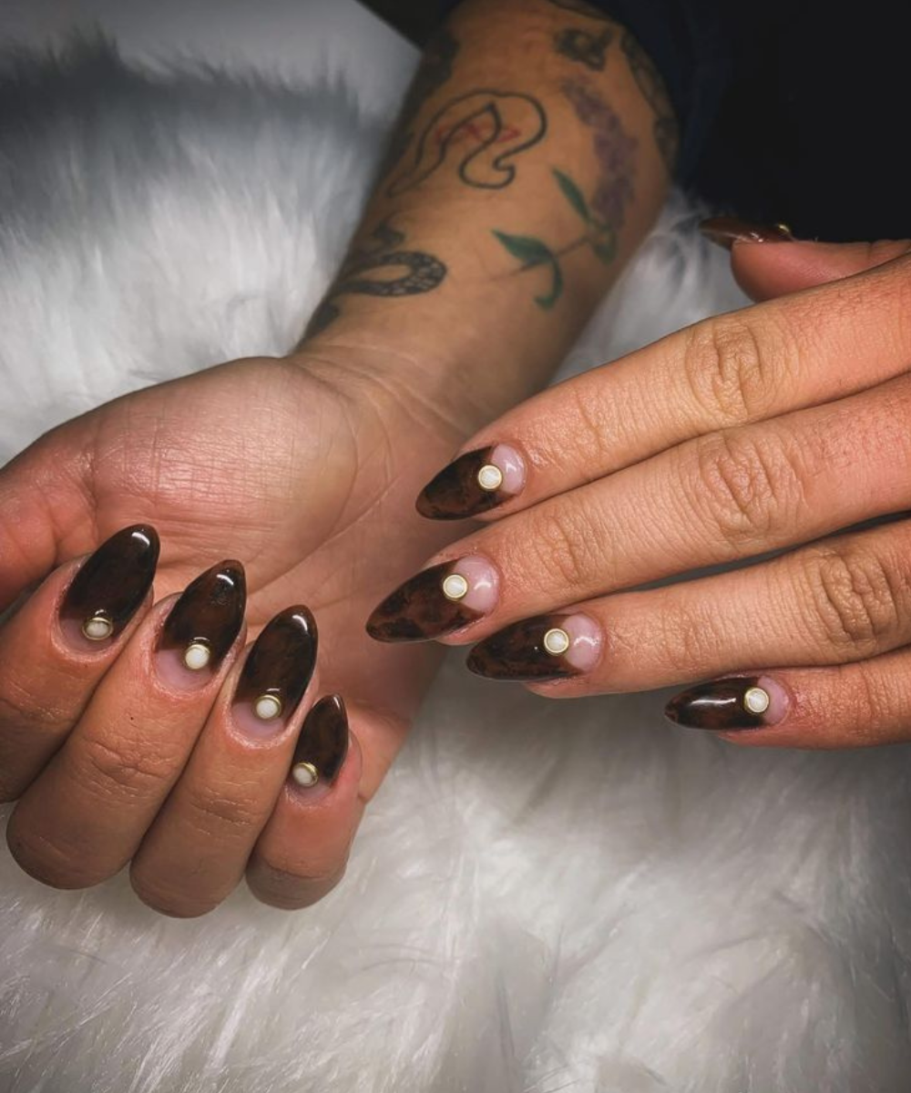 Esmaltação polêmica com unhas longas em formato amendoado, esmalte marrom translúcido e detalhe circular branco, estilo