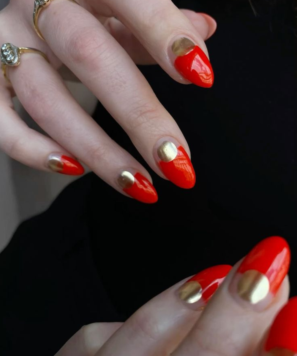 Esmaltação polêmica com unhas longas em formato amendoado, combinando vermelho vibrante e detalhes dourados metálicos,