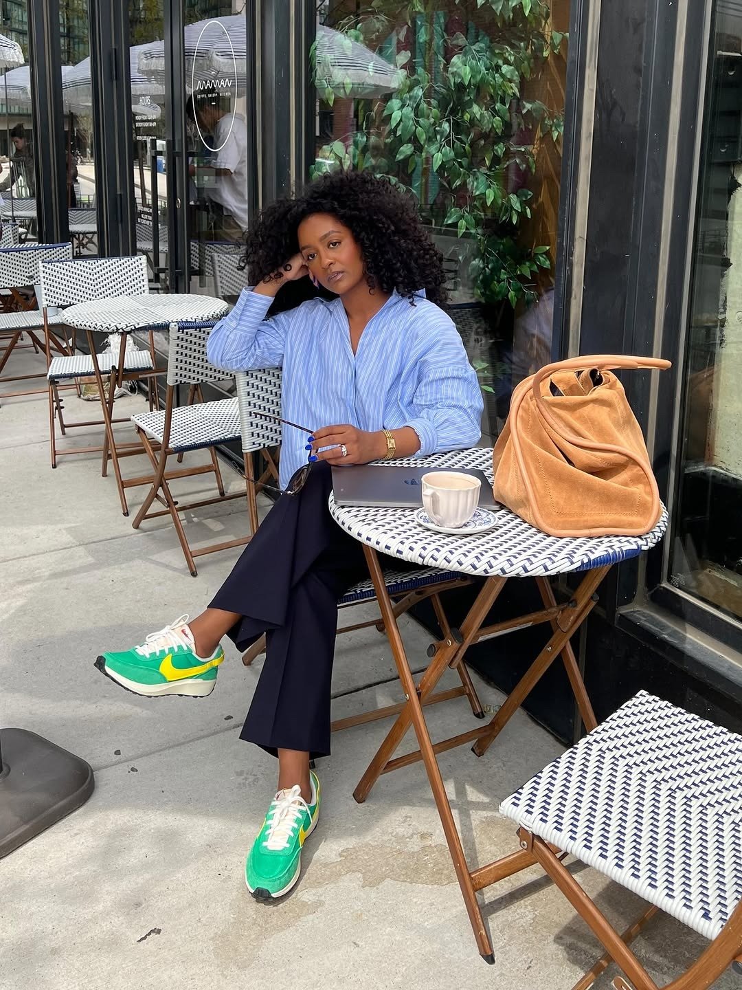 Mulher sentada em café urbano usa tênis da Nike verde e amarelo com calça preta e camisa azul listrada, em pose pensativa e