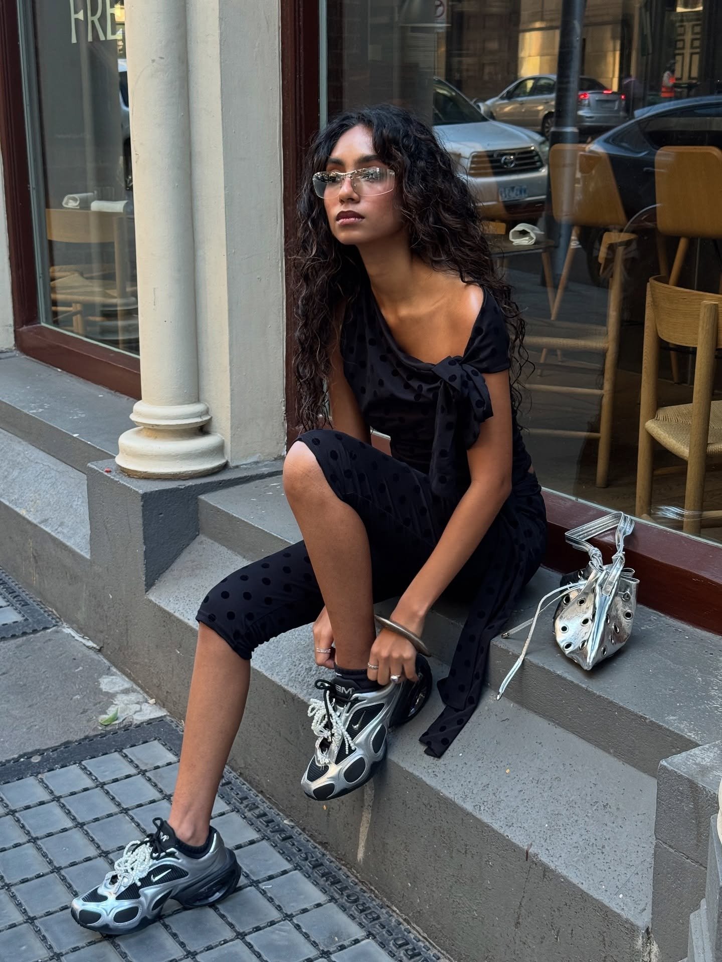 Modelo senta casualmente em degrau urbano usando vestido midi preto de poás com laço no ombro e tênis da Nike prata e preto