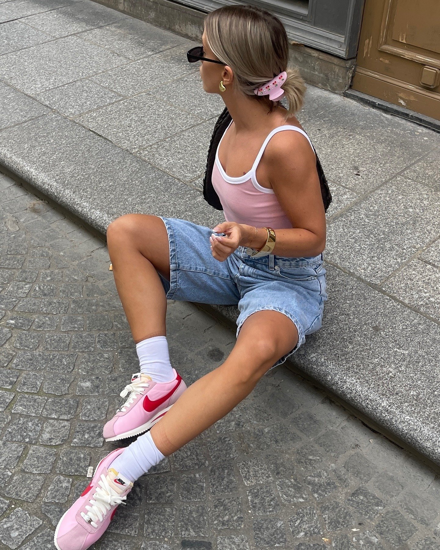 Mulher sentada na calçada usa tênis da Nike rosa com detalhe vermelho, bermuda jeans e regata rosa, estilo casual e urbano.