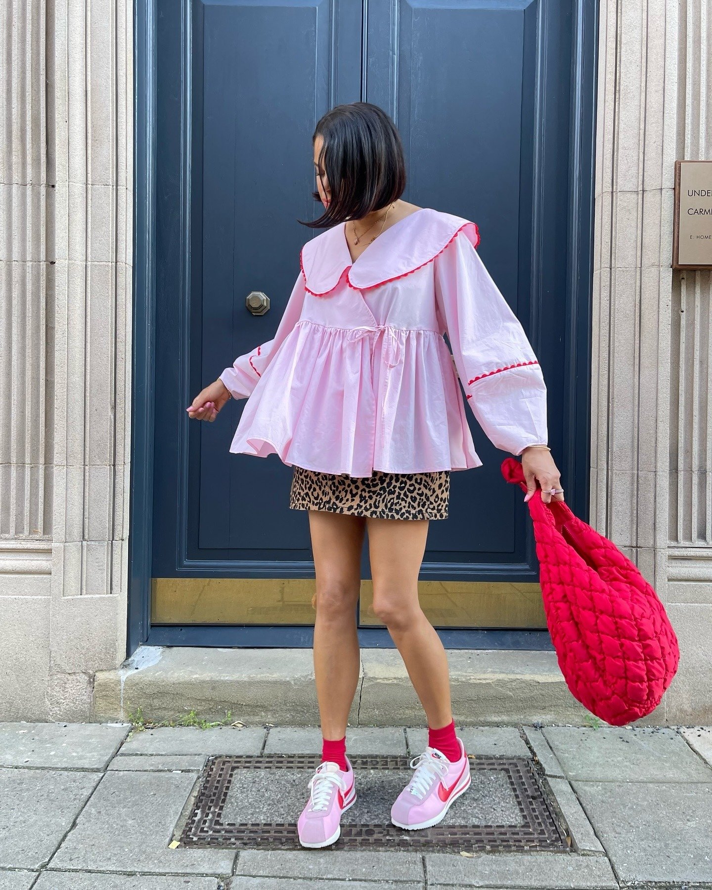 Modelo posa em frente a porta azul usando tênis da Nike rosa com detalhes vermelhos, saia animal print e blusa ampla rosa