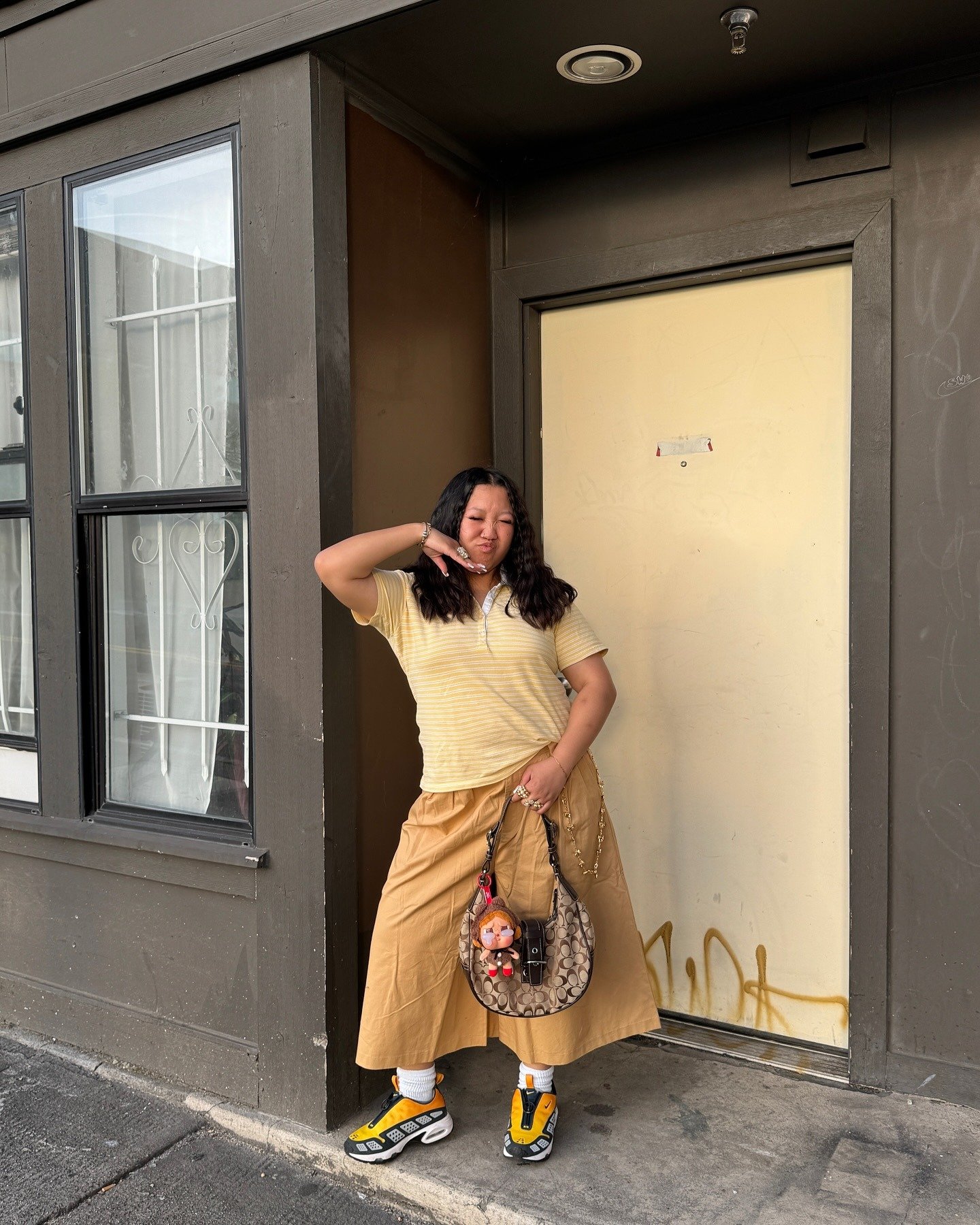 Modelo posa com camiseta polo listrada amarela, saia midi caramelo e tênis da Nike amarelo com detalhes em preto e branco