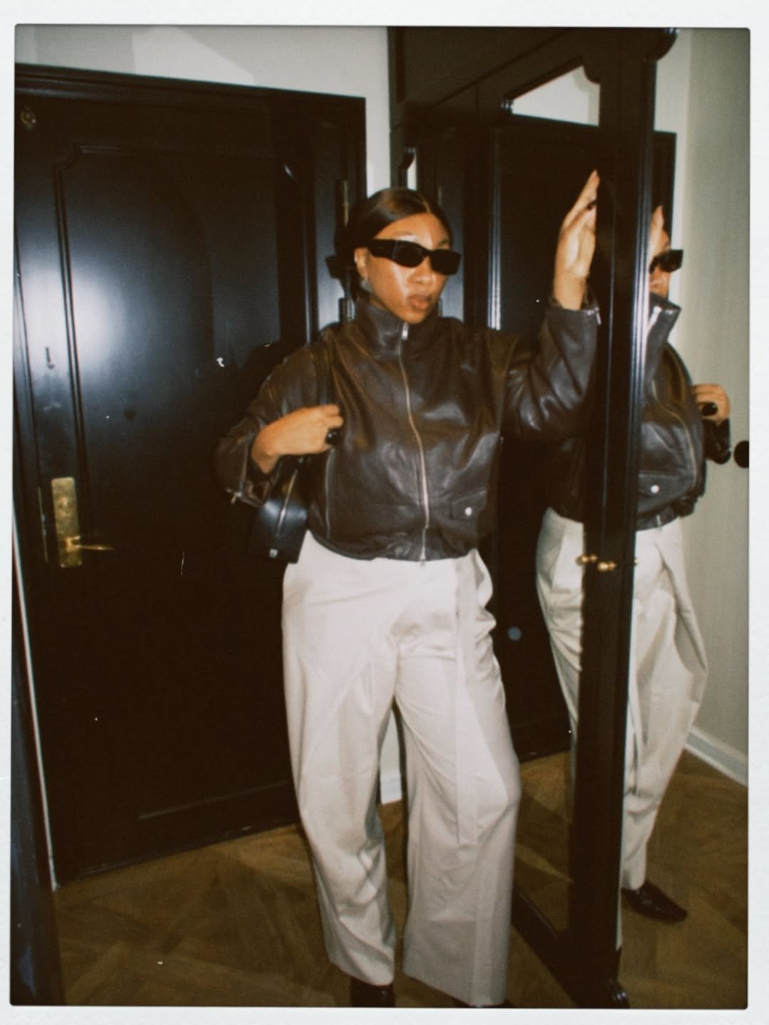 Looks com calça wide leg bege e jaqueta de couro preta; modelo posa de óculos escuros em clima urbano sofisticado.