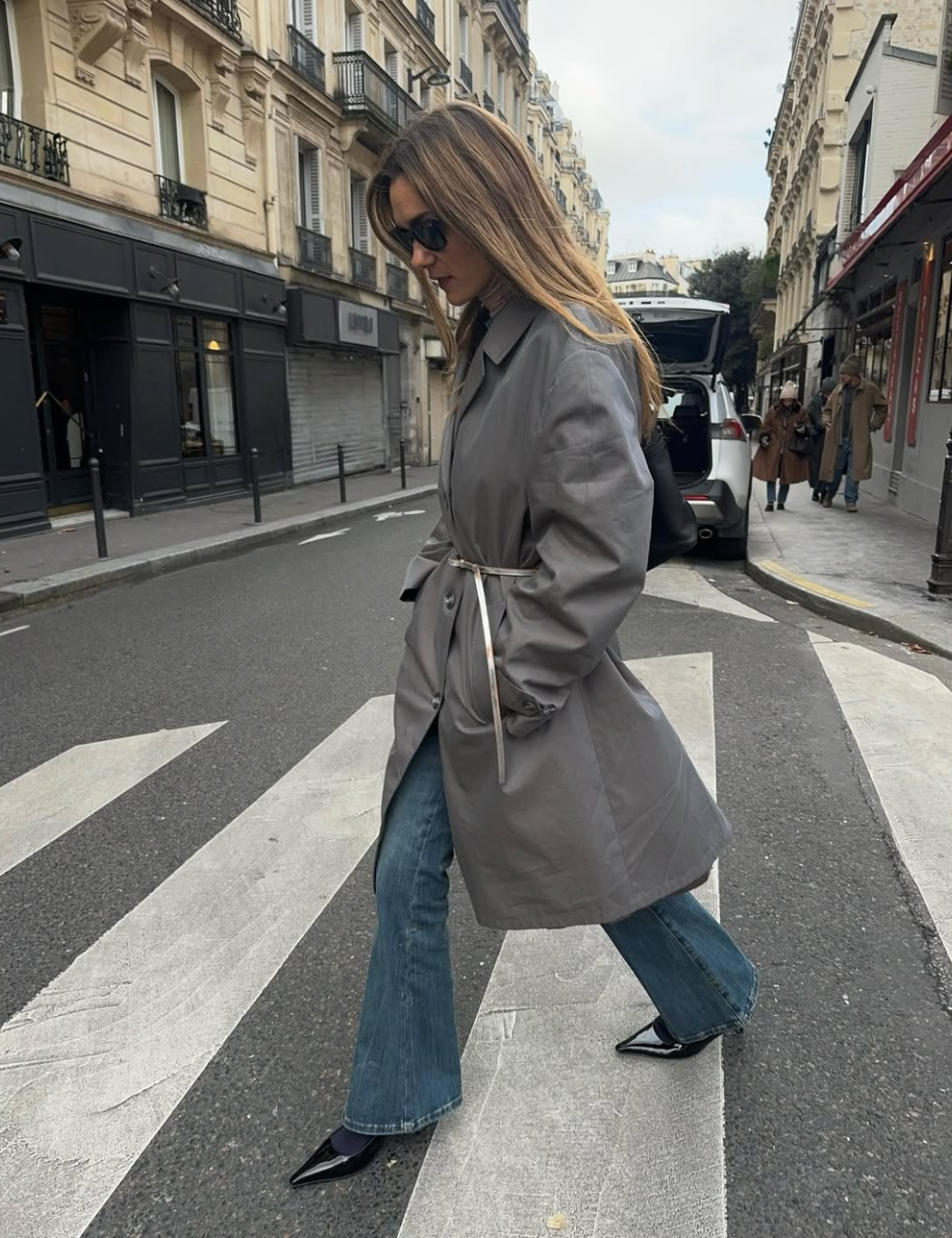 Mulher andando na rua mostra o que é calça boot cut ao combiná-la com trench coat e bota preta.