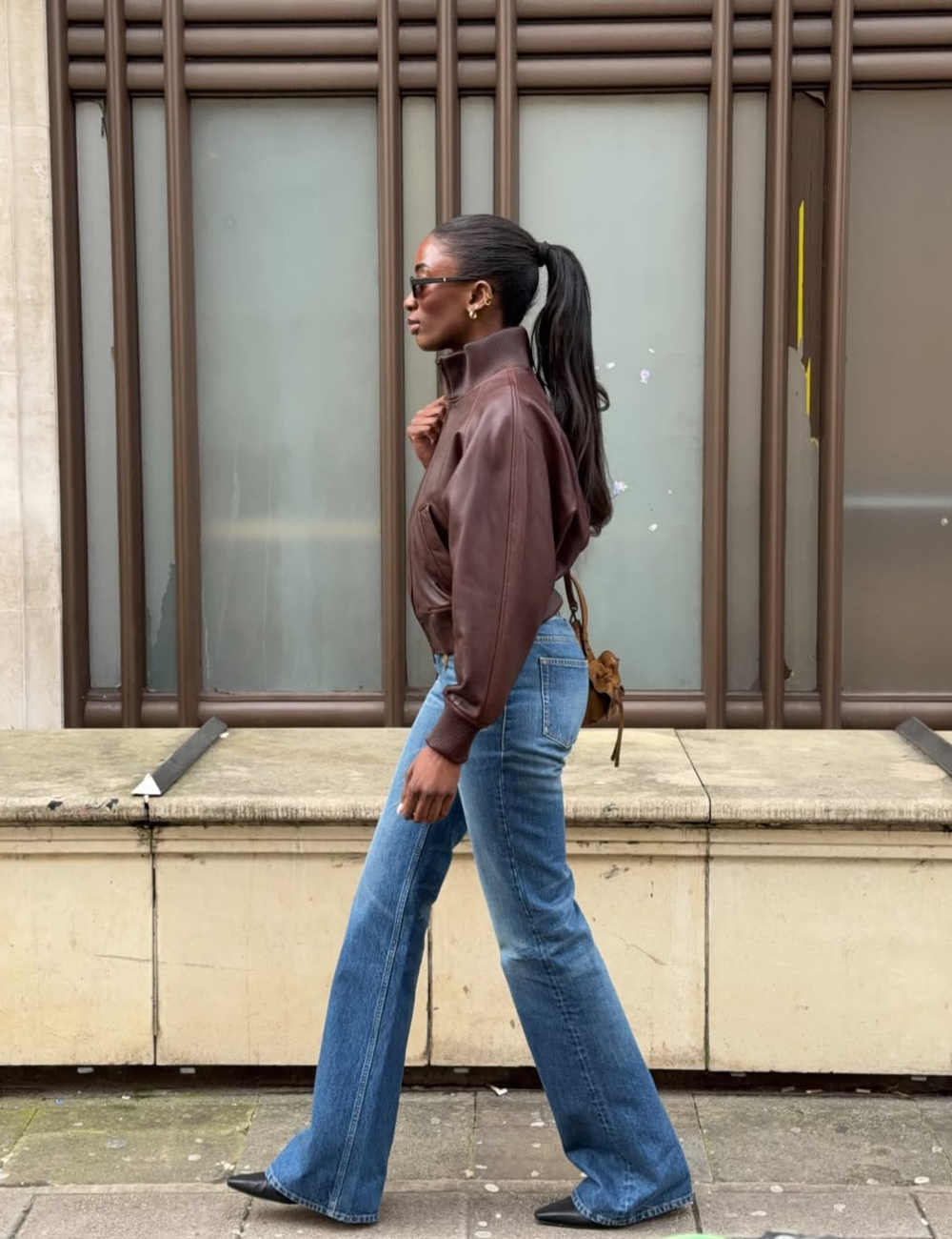Mulher andando na rua usando rabo de cavalo, calça jeans estilo boot cut, jaqueta de couro marrom e sapatos de bico fino.