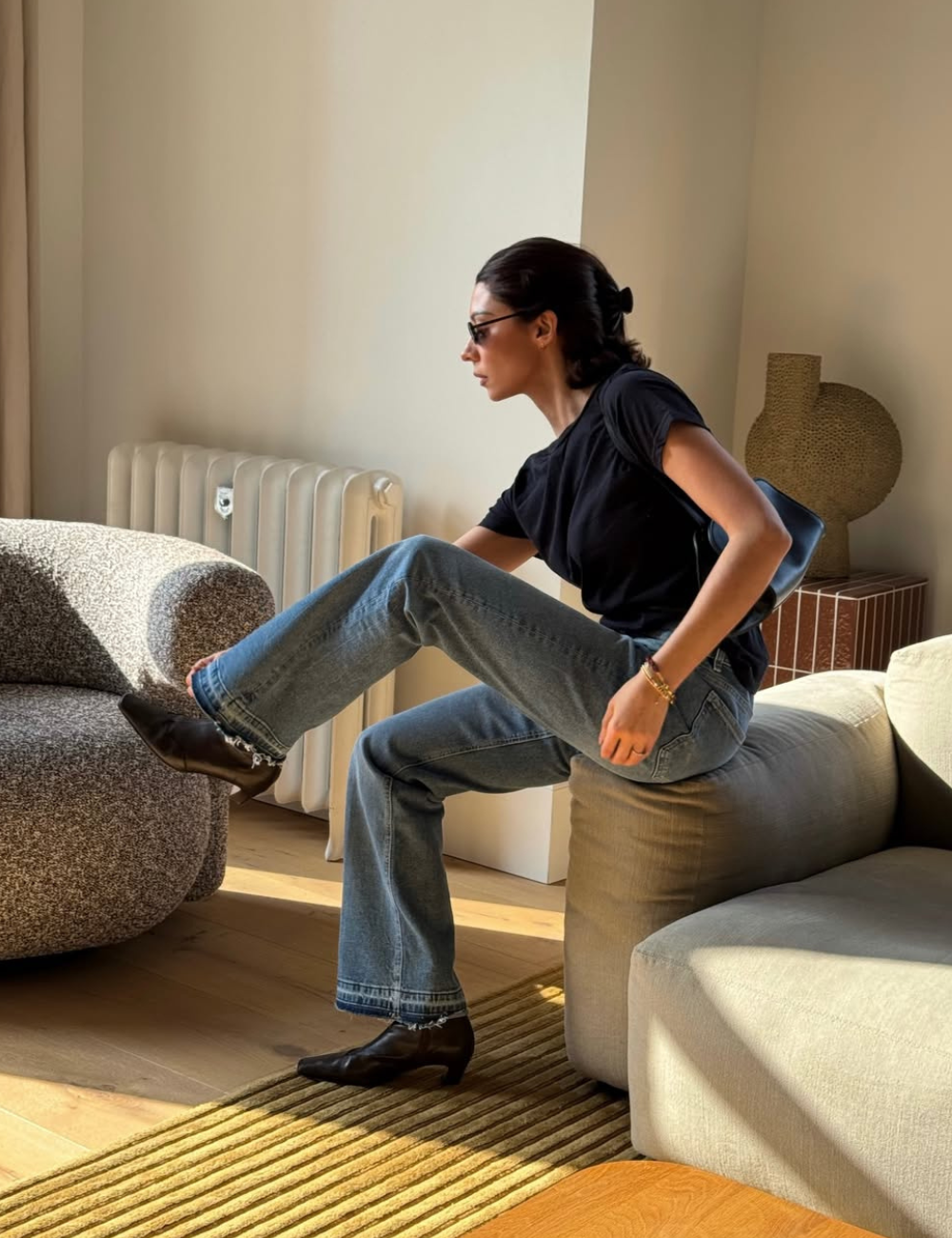 Mulher em ambiente aconchegante, sentada no sofá usando camiseta preta básica, calça boot cut e botas.