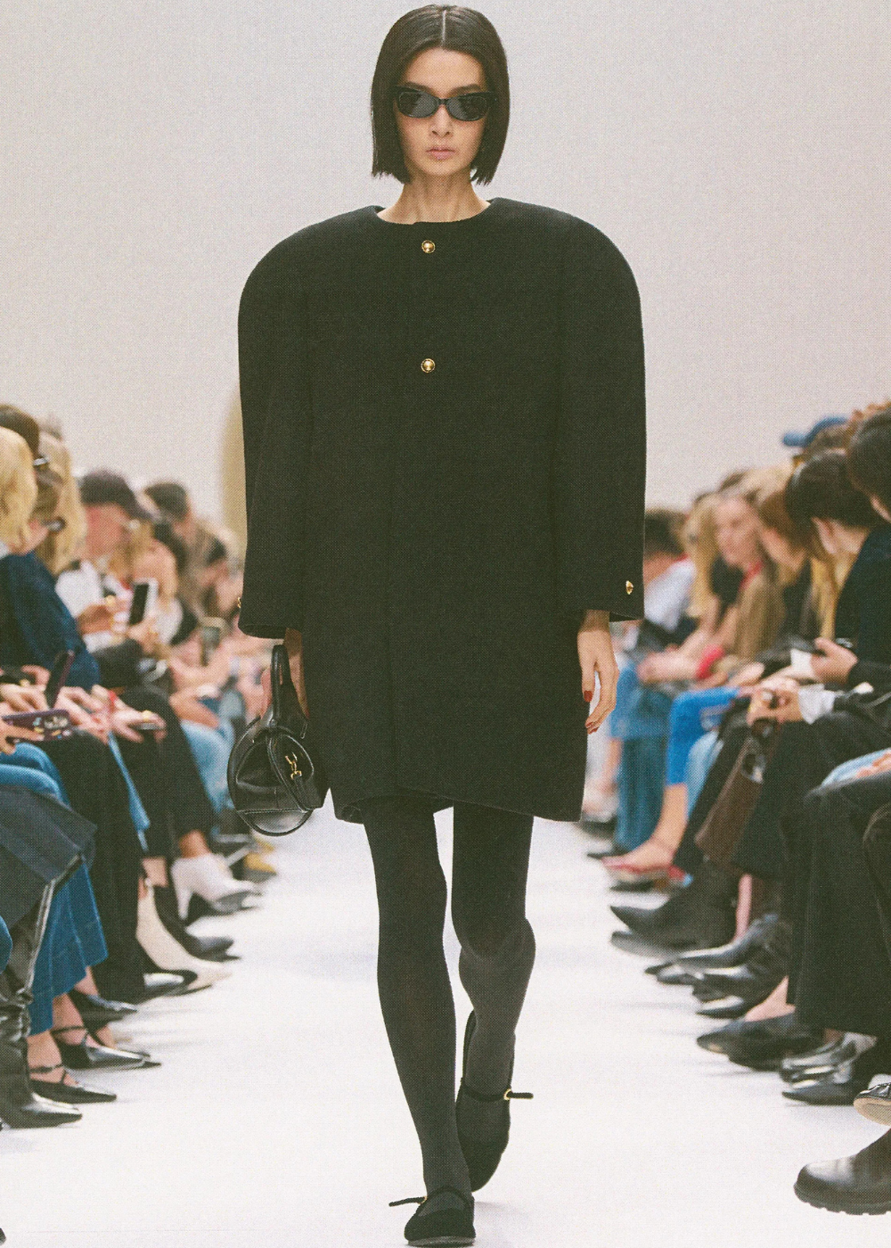 Outono/Inverno 2026: modelo desfila de preto com vestido amplo estruturado, óculos escuros e clima sóbrio na passarela.