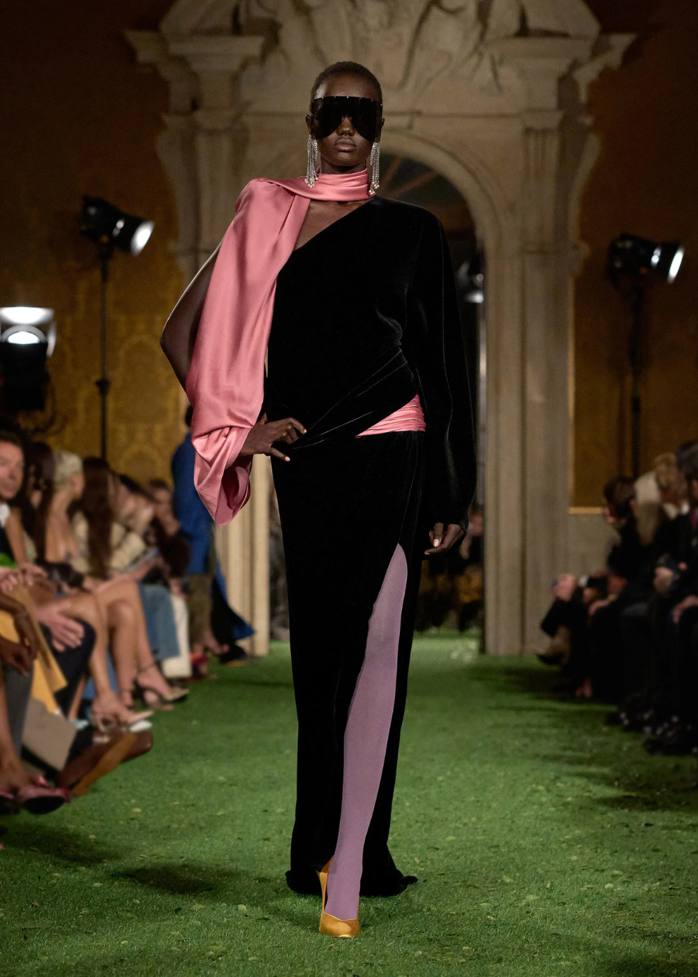 Outono/Inverno 2026: modelo desfila vestido assimétrico de veludo preto e drapeado rosa em passarela clássica, clima