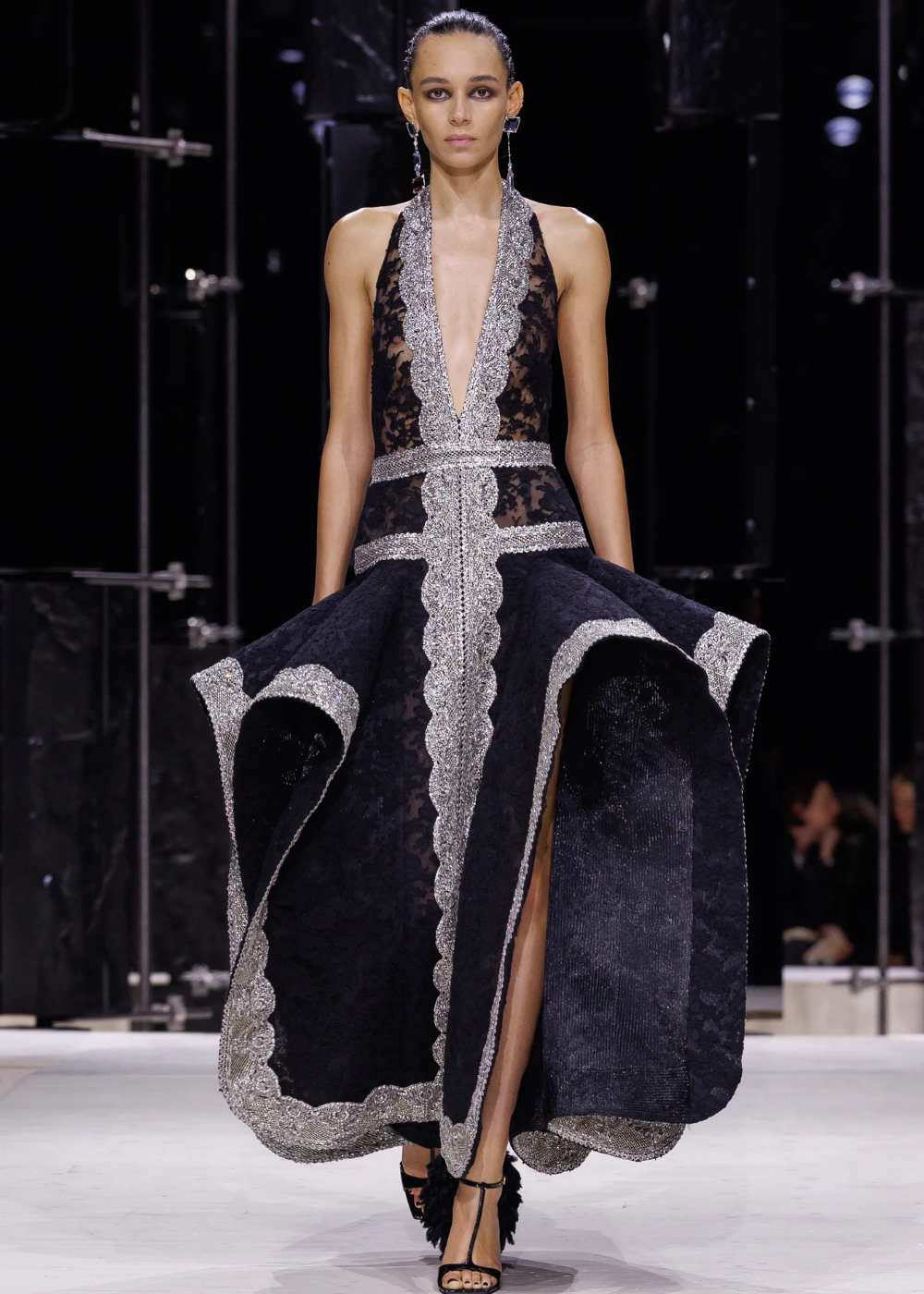 Outono/Inverno 2026: modelo posa na passarela com vestido preto rendado, decote profundo e bordas prateadas, em clima