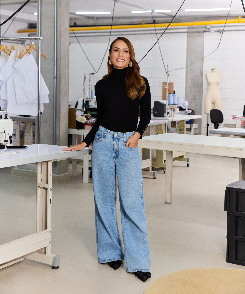PatBo: modelo posa sorrindo em gola alta preta e jeans wide leg azul-claro em ateliê, com elegância contemporânea.