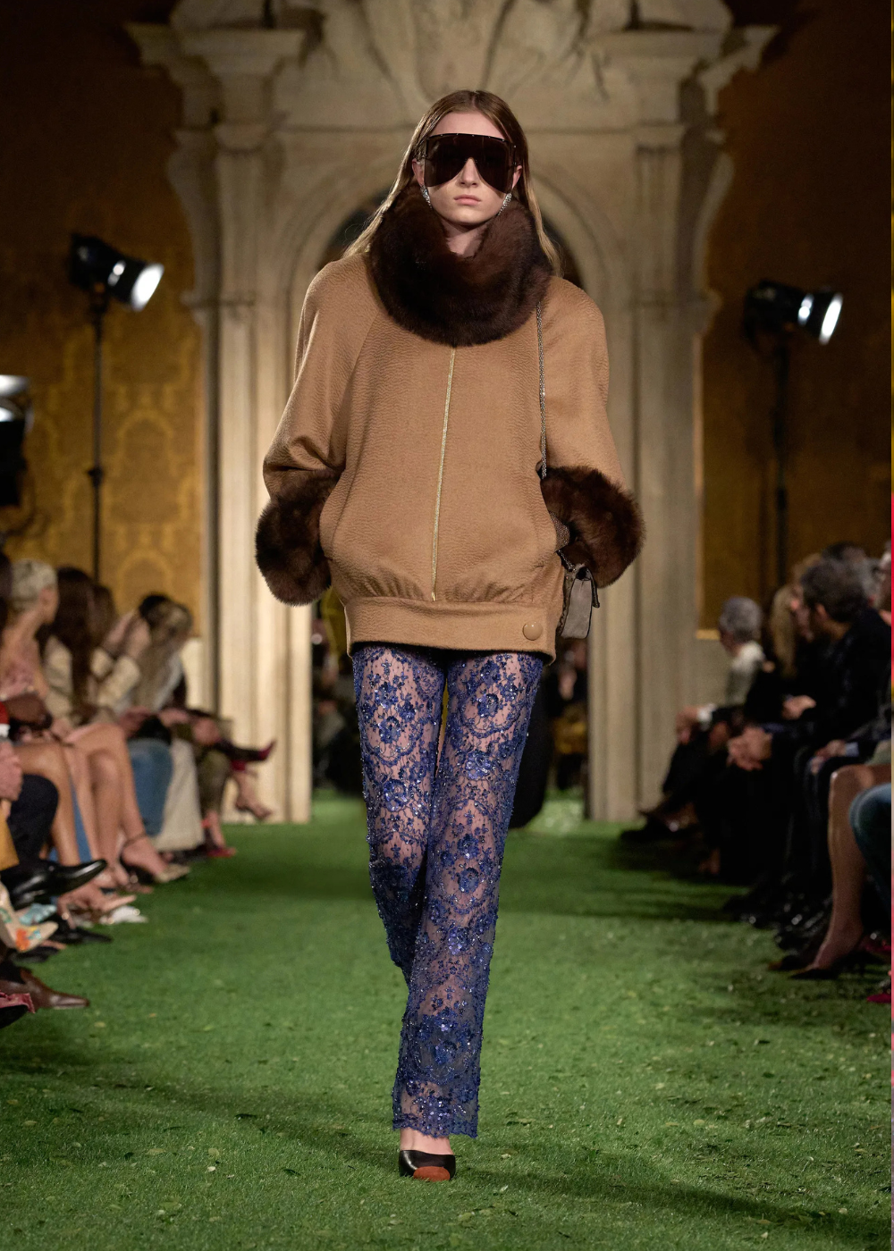 Outono/Inverno 2026: modelo desfila com blusão camel de pele e renda azul, em passarela clássica de clima sofisticado