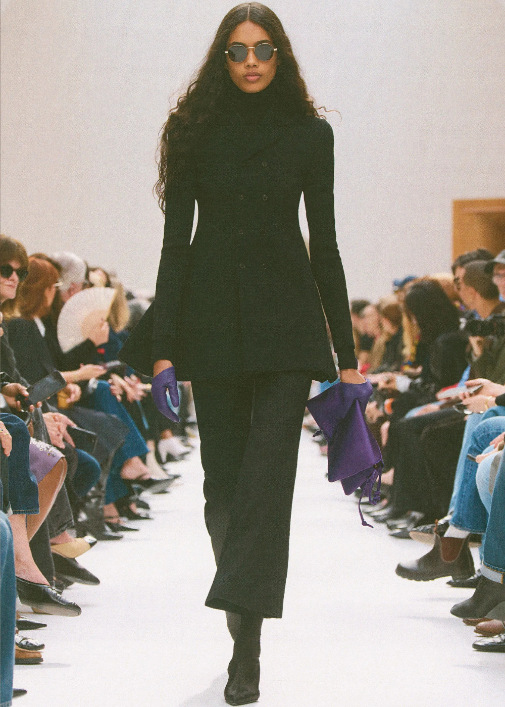 Outono/Inverno 2026: modelo desfila de preto com casaco acinturado, calça ampla, óculos redondos e luvas roxas.