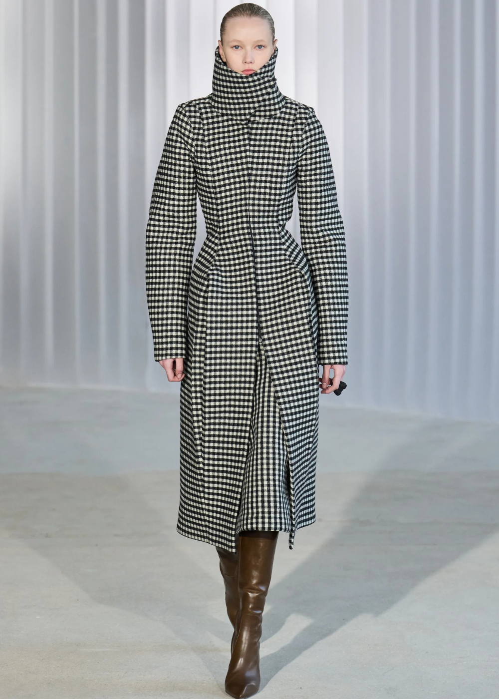 Outono/Inverno 2026: modelo posa com casaco xadrez acinturado de gola alta e botas marrons em clima sóbrio.