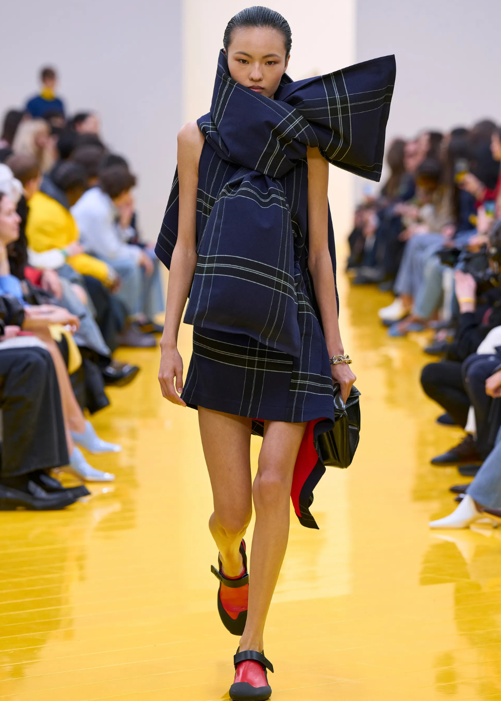 Outono/Inverno 2026: modelo desfila vestido xadrez oversized azul-marinho, laço escultórico e sapatos vermelho e preto.