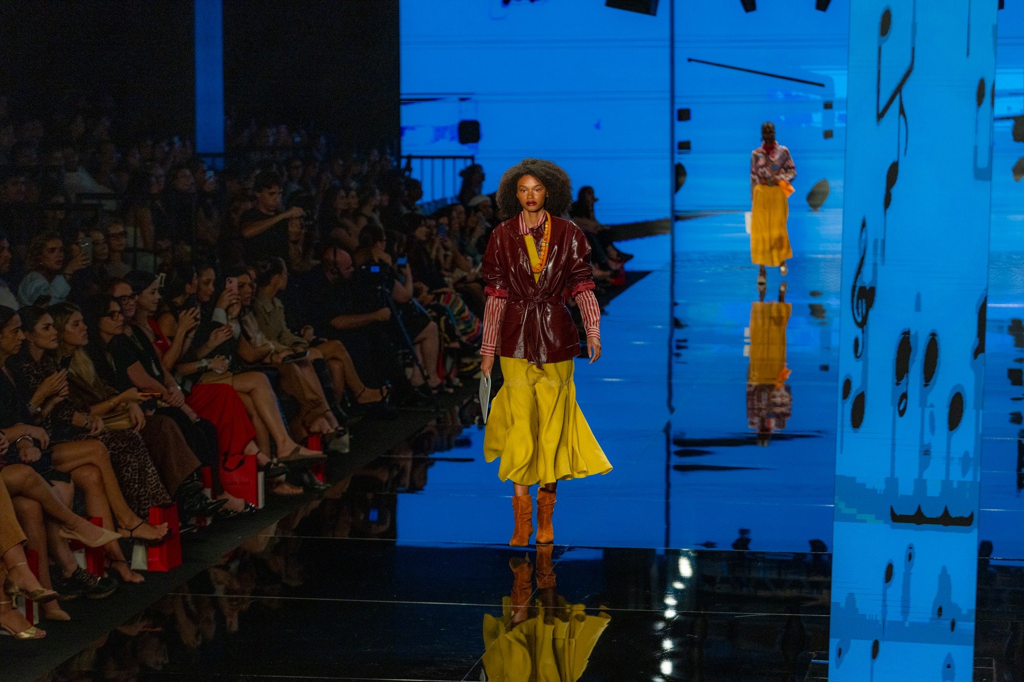 Moda inverno 2026: modelo desfila com casaco vinho brilhante, saia amarela ampla e clima sofisticado.