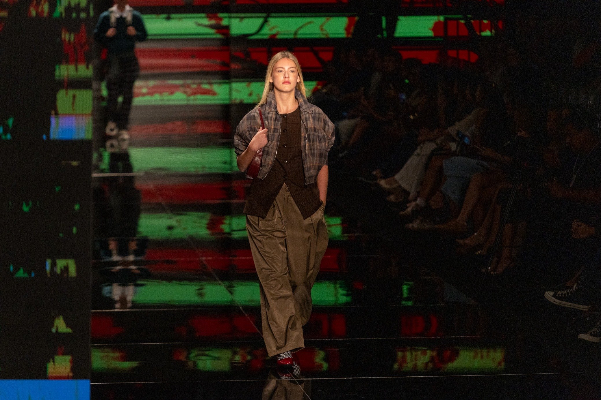 Moda inverno 2026: modelo desfila com jaqueta xadrez curta, colete escuro e calça ampla cáqui em passarela dramática.