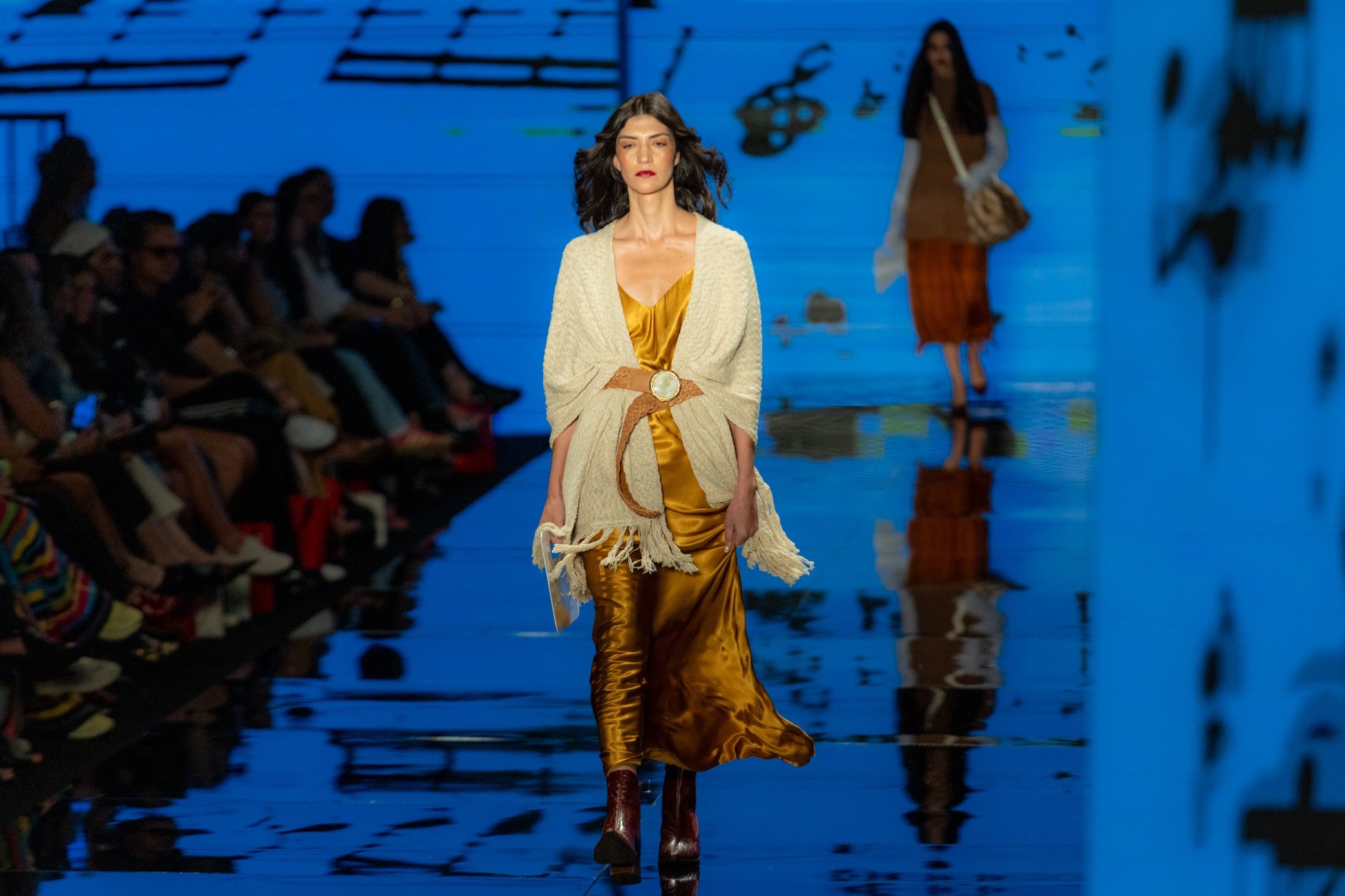 Moda inverno 2026: modelo desfila com vestido acetinado dourado, xale cru com franjas e clima sofisticado na passarela azul