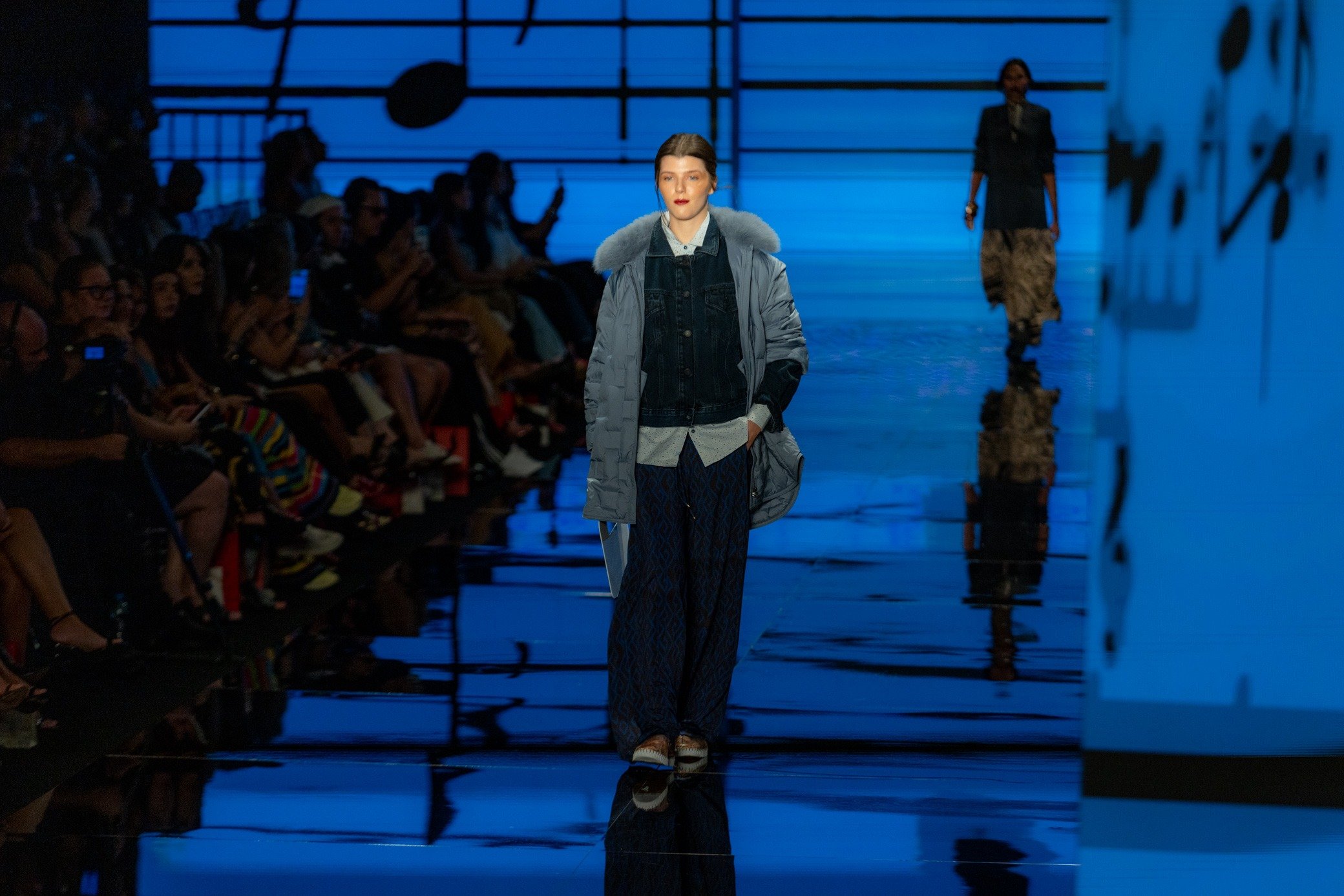 Moda inverno 2026: modelo desfila com jaqueta acolchoada azul, jeans e calça ampla em passarela azul de clima sóbrio.