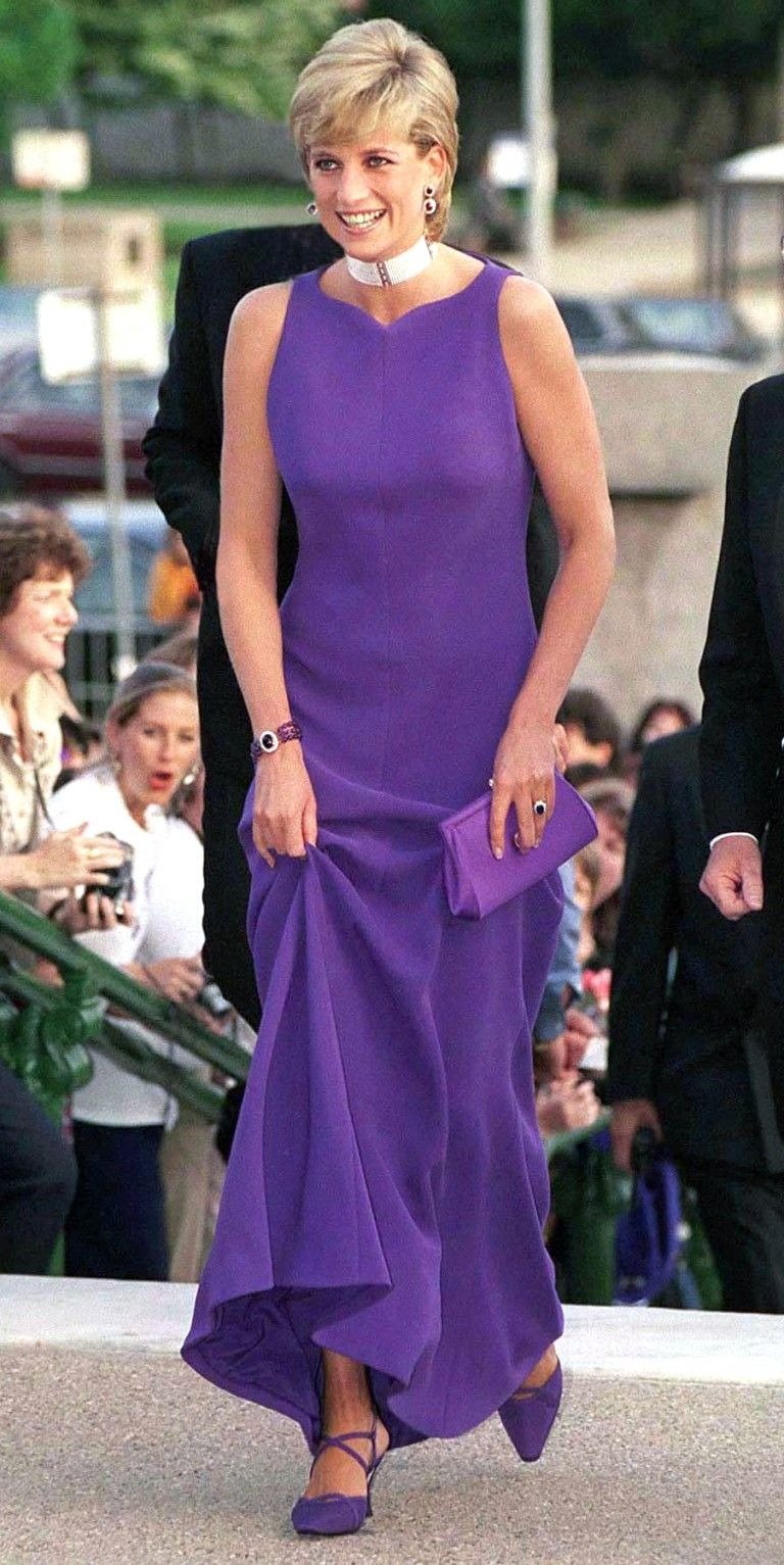 Modelo posa sorridente com vestido longo Versace roxo de modelagem reta, tecido fluido, clutch e sapatos combinando, em