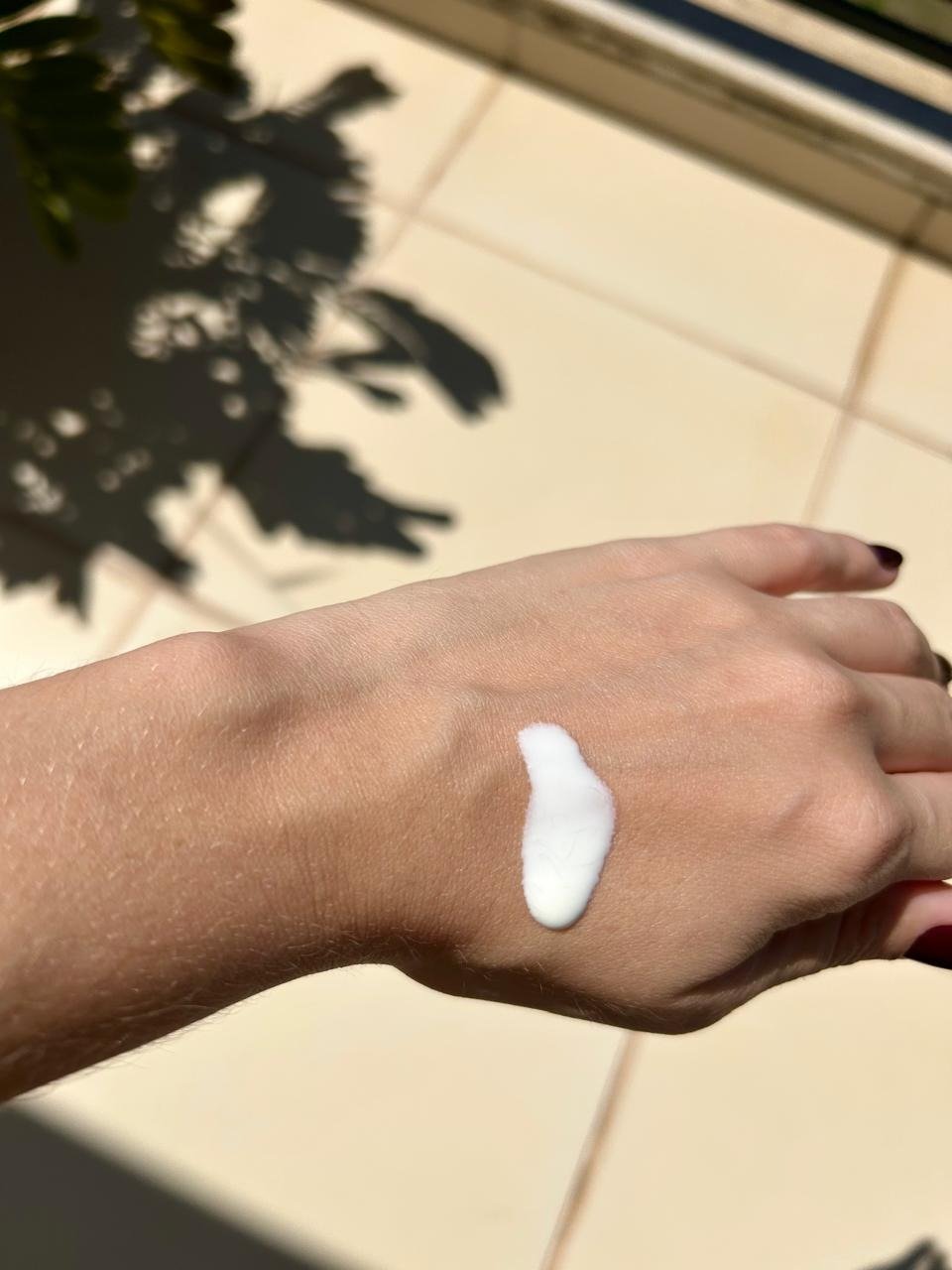 Protetor solar hidratante da The Ordinary em textura cremosa branca sobre a mão, sob sol forte e sombra de folhas.