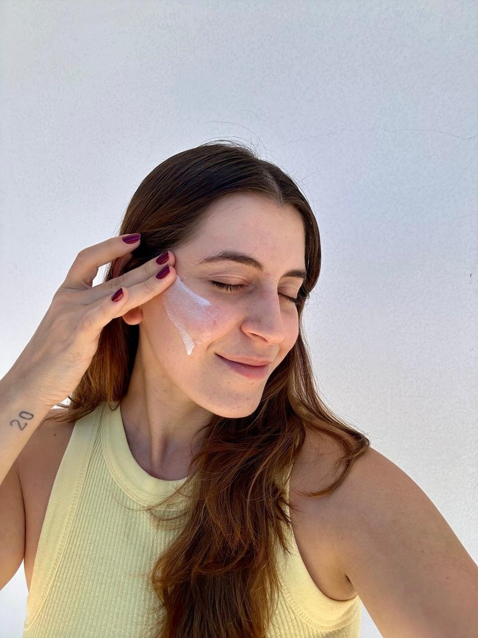 Modelo posa com protetor solar hidratante da The Ordinary no rosto, regata canelada amarela e clima leve.