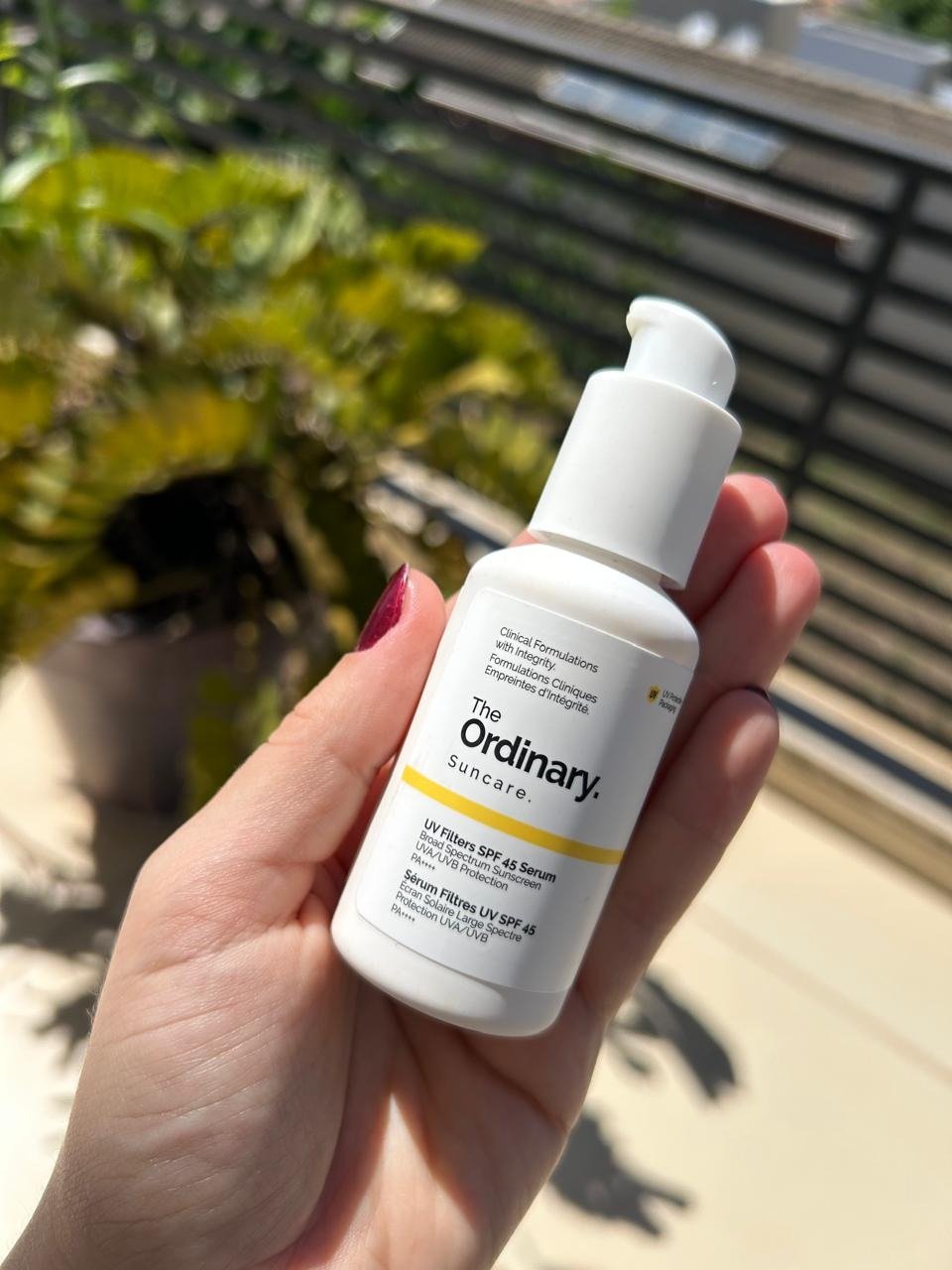 Mão segura protetor solar hidratante da The Ordinary branco ao sol, com fundo verde desfocado; Texto: The Ordinary.