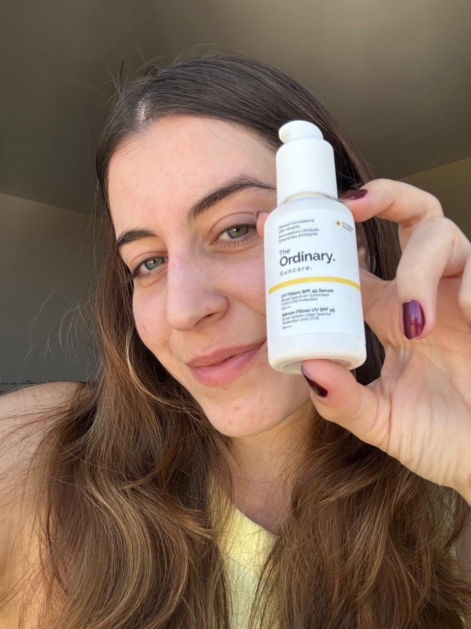 Modelo posa com protetor solar hidratante da The Ordinary em frasco branco; top amarelo e clima leve.
