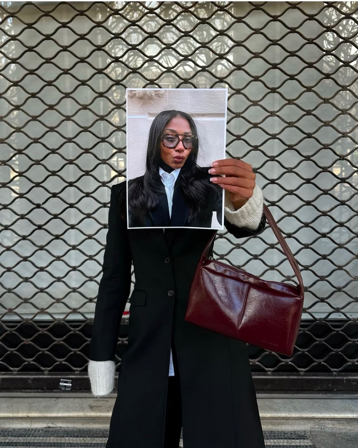 Rede social de fotos com modelo posa de casaco preto, punhos de tricô e bolsa vinho diante de grade urbana, clima cool.