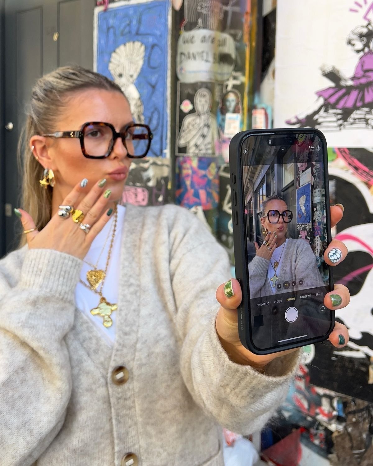 Modelo posa para rede social de fotos com cardigan cinza, óculos maxi e joias douradas diante de mural urbano colorido.