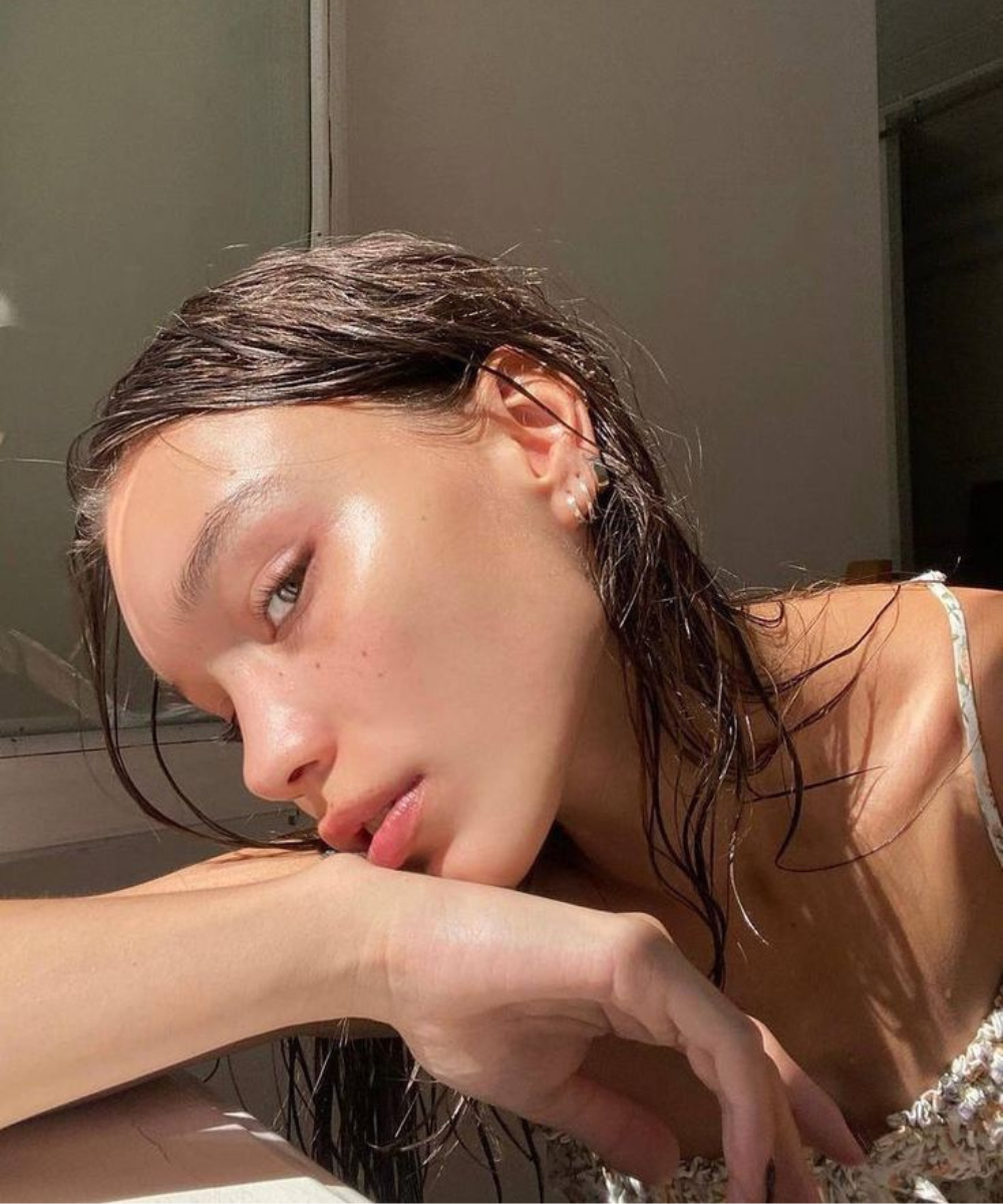 Modelo posa com expressão serena, pele luminosa e natural, cabelos molhados e vestido de alças finas em tom neutro, evocando