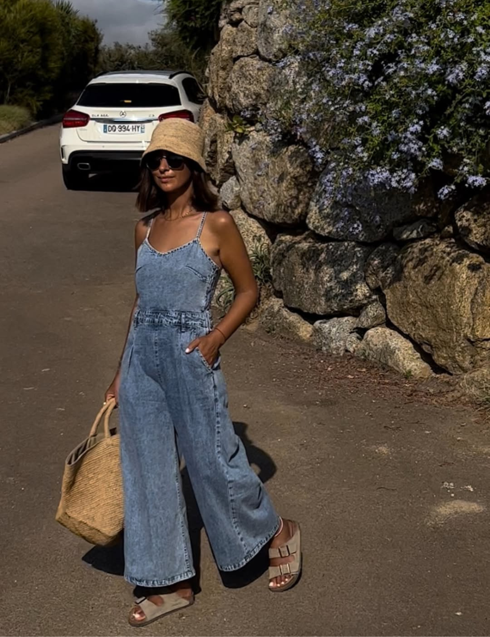 Mulher em pé usando macacão jeans, chapéu de palha e sandália de fivela estilosa em cenário praiano.