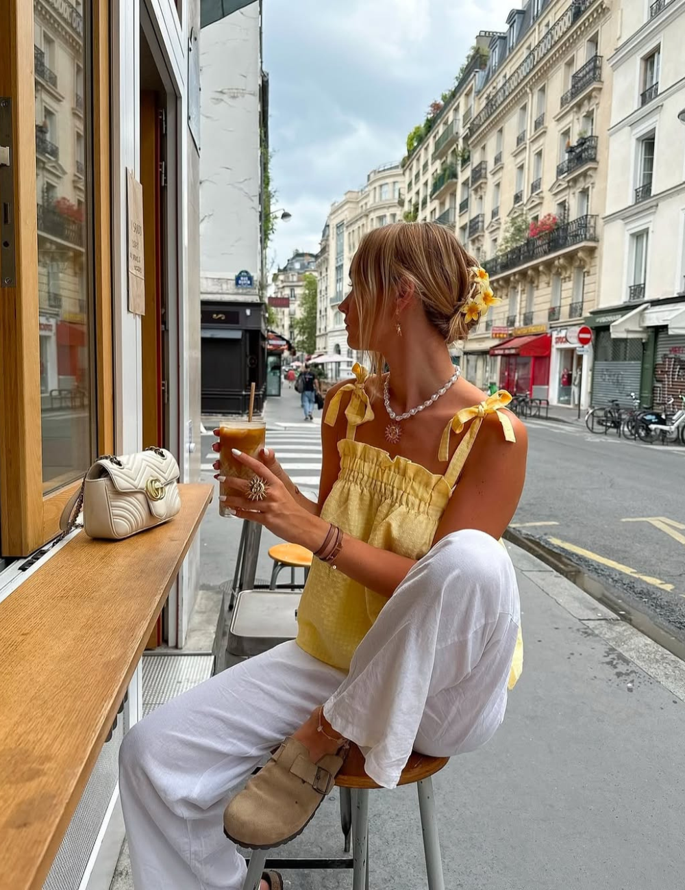 Modelo sentada usando calças jeans brancas, blusa amarela e clog de camurça enquanto bebe um café.