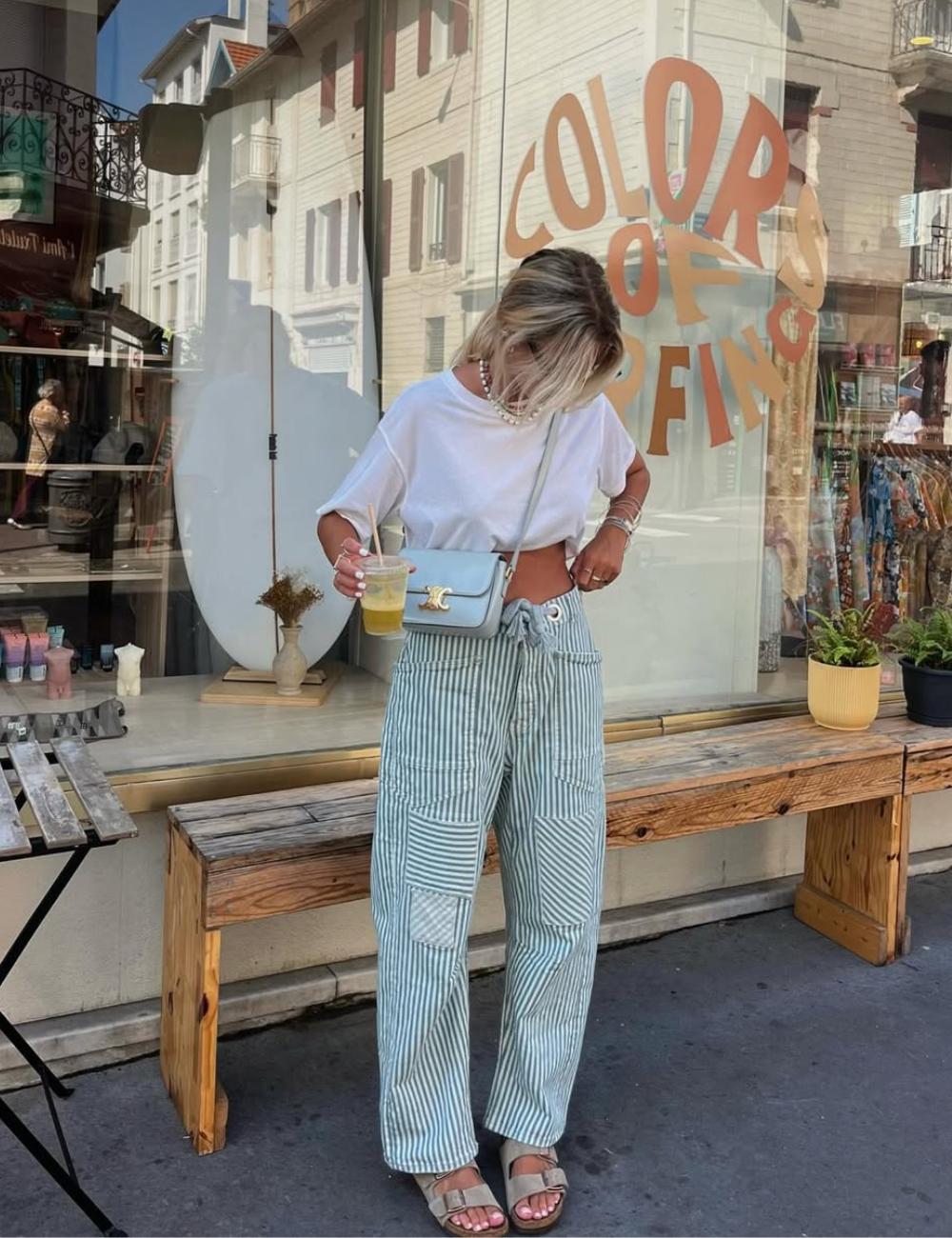 Mulher de calça listrada em pé, arrumando a blusa branca enquanto usa sandália de fivela estilosa em clima leve.