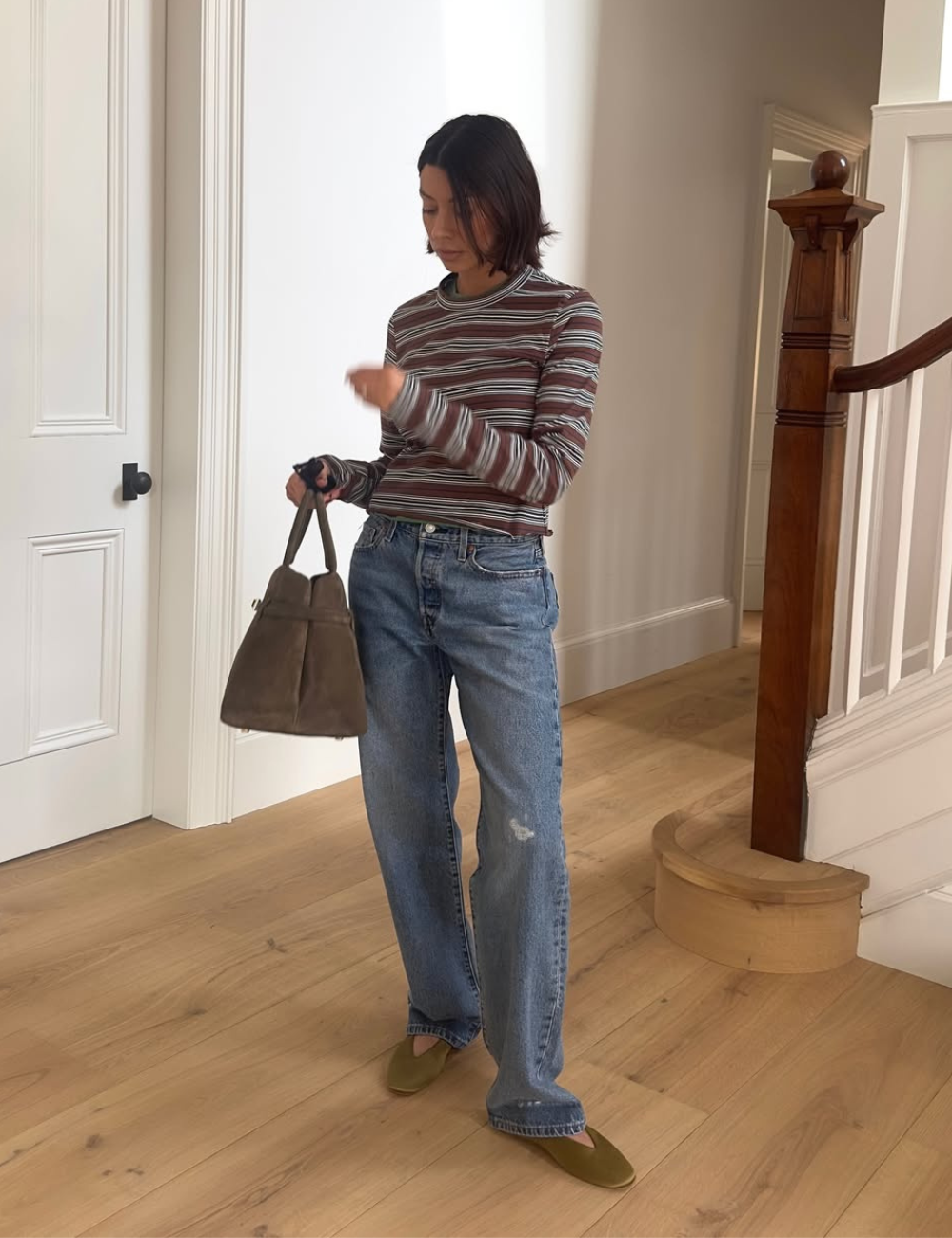 Modelo posa com sapatilha amendoada verde-oliva, blusa listrada justa, jeans amplo e bolsa marrom em hall sereno.