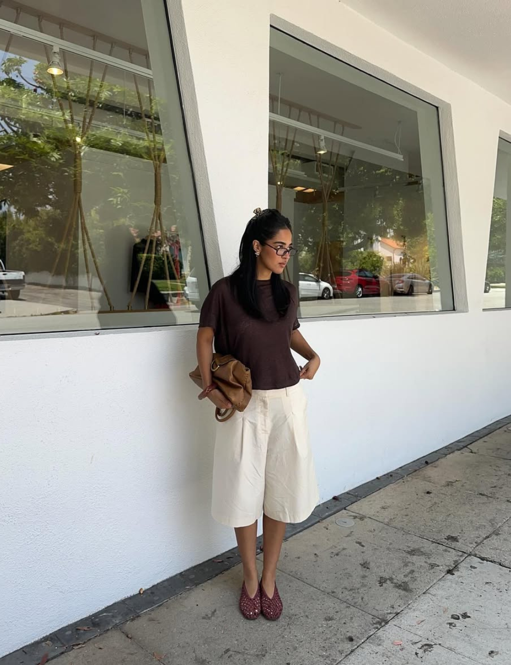 Modelo posa com sapatilha amendoada vazada vinho, blusa marrom, bermuda off-white e bolsa camel.