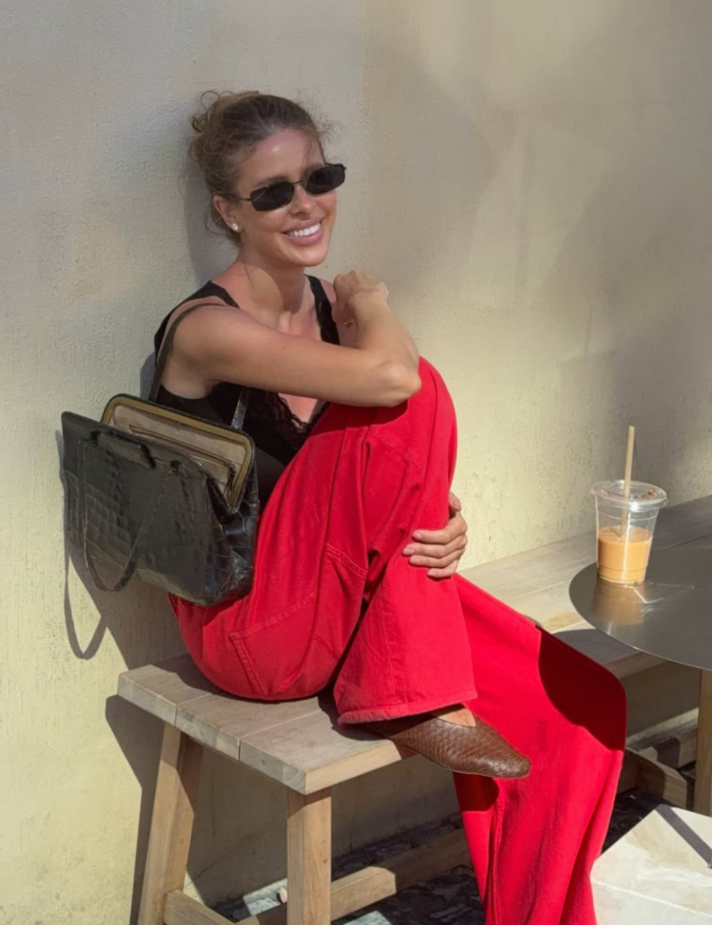 Modelo posa sorrindo com sapatilha amendoada marrom, calça vermelha ampla, regata preta e bolsa croco ao sol.