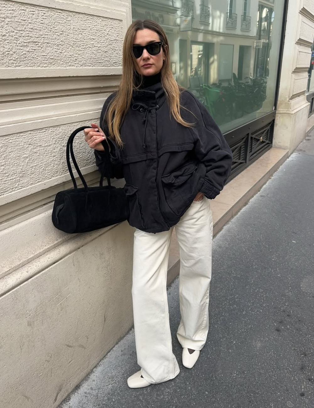 Modelo posa na calçada com sapatilha amendoada off-white, jaqueta preta oversized, calça ampla e bolsa de camurça.