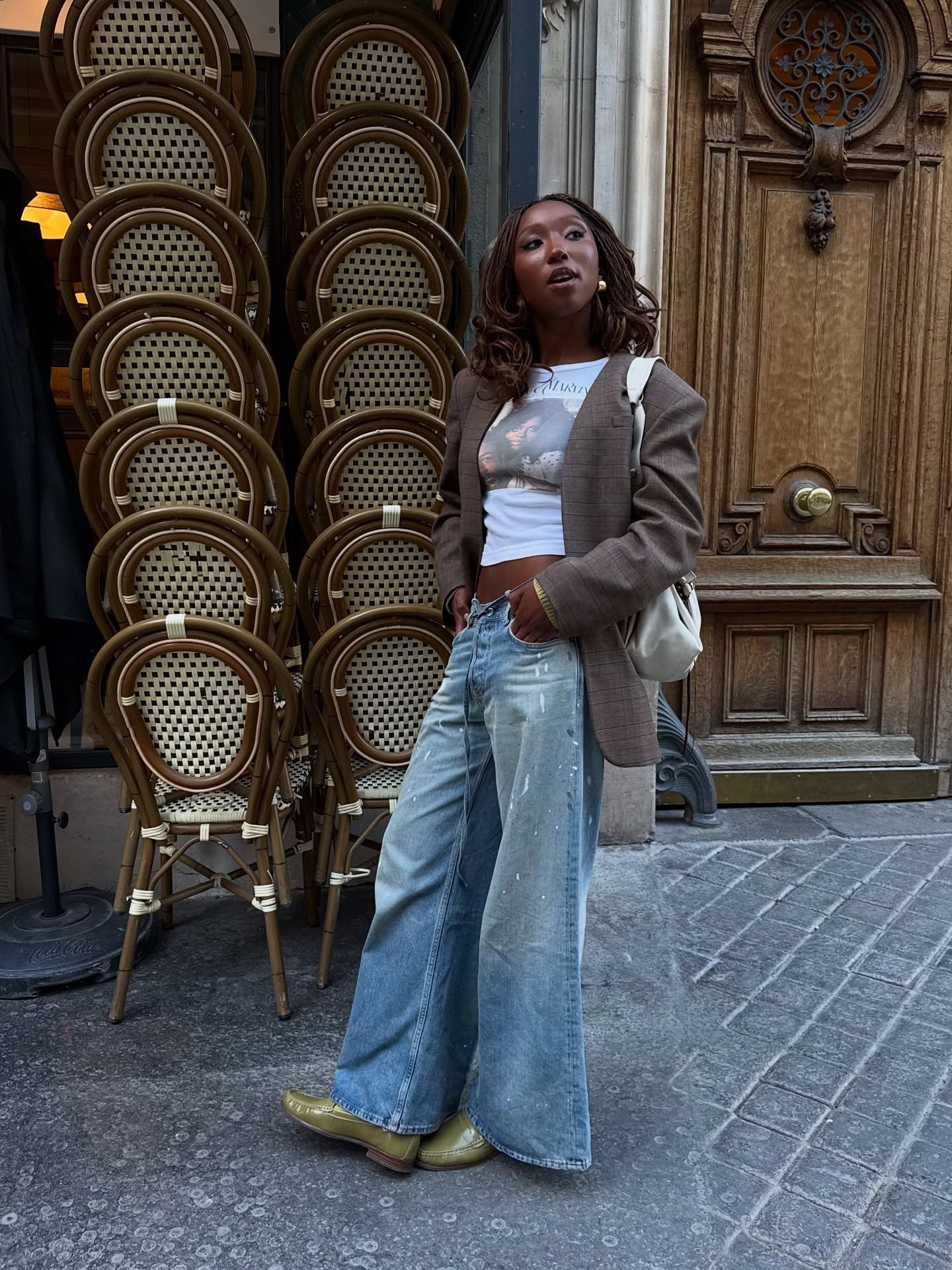 Modelo posa com sapatos coloridos verde-oliva, blazer xadrez, jeans amplo e bolsa branca em rua elegante.