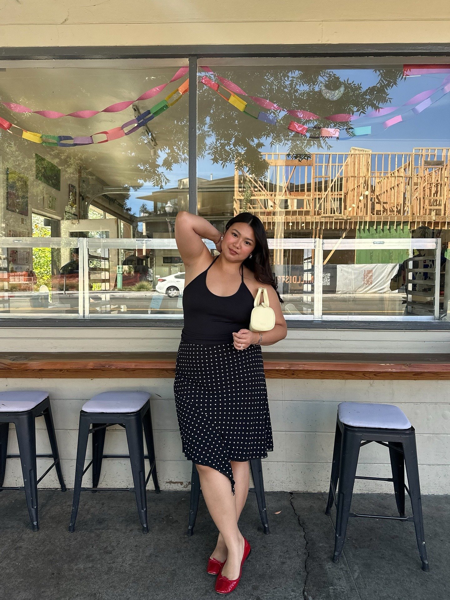 Modelo posa com sapatos coloridos vermelhos, regata preta e saia midi de poás diante de vitrine festiva, clima leve.