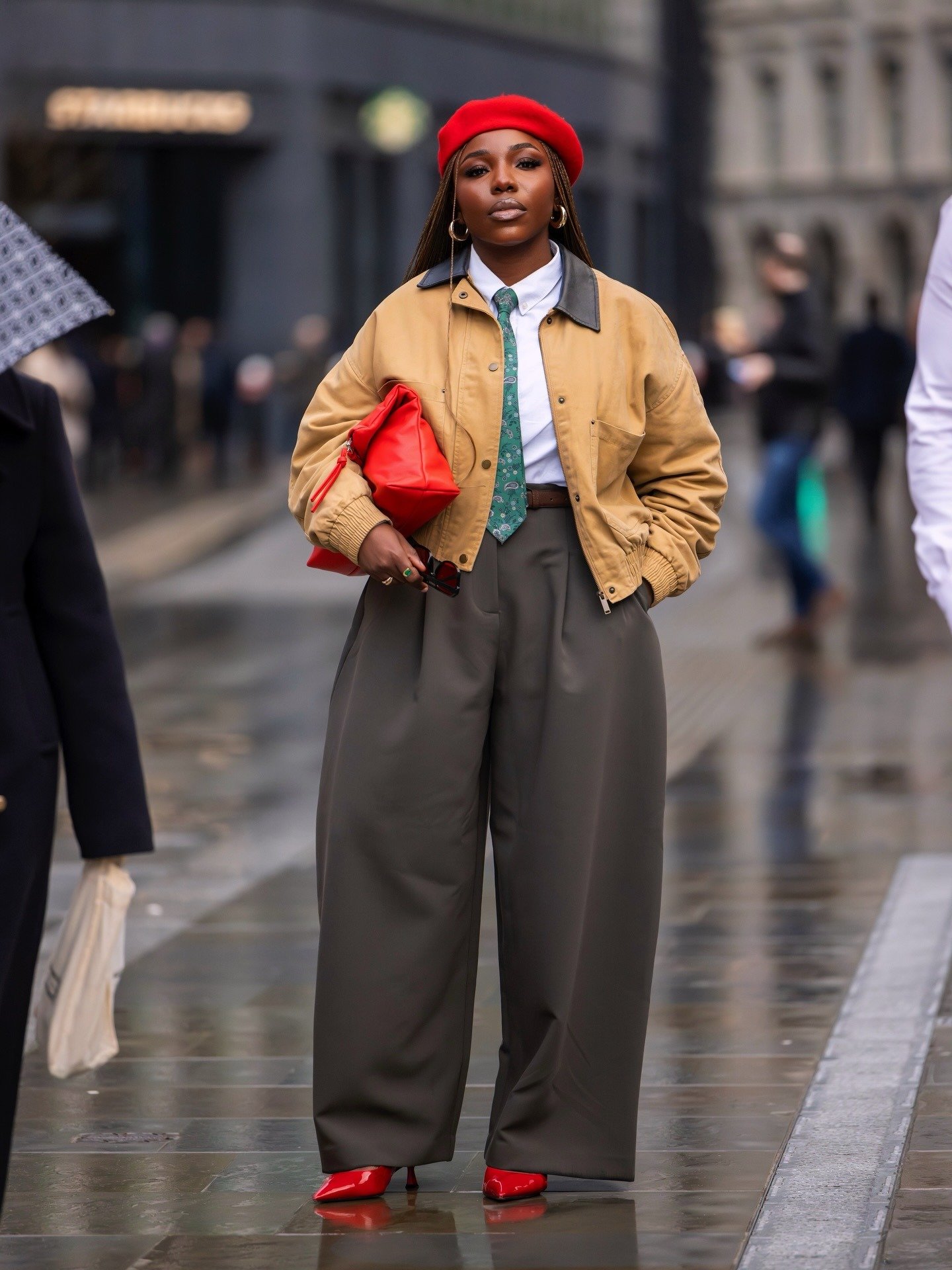 Modelo posa na rua com sapato colorido vermelho, boina, jaqueta bege, calça ampla e bolsa vibrante.