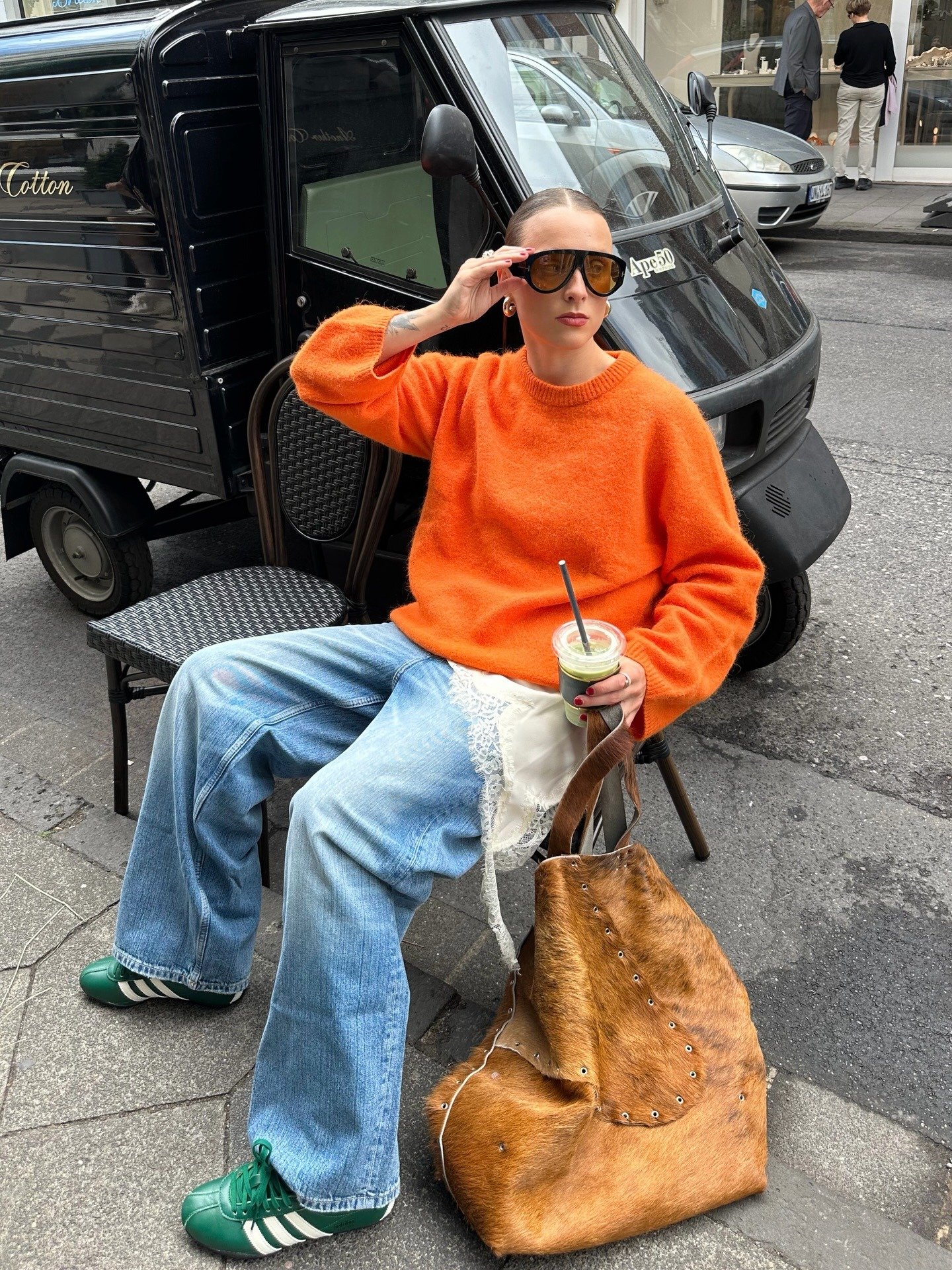 Modelo posa sentada com sapatos coloridos verdes, tricô laranja, jeans amplo e bolsa de pelo na calçada urbana.