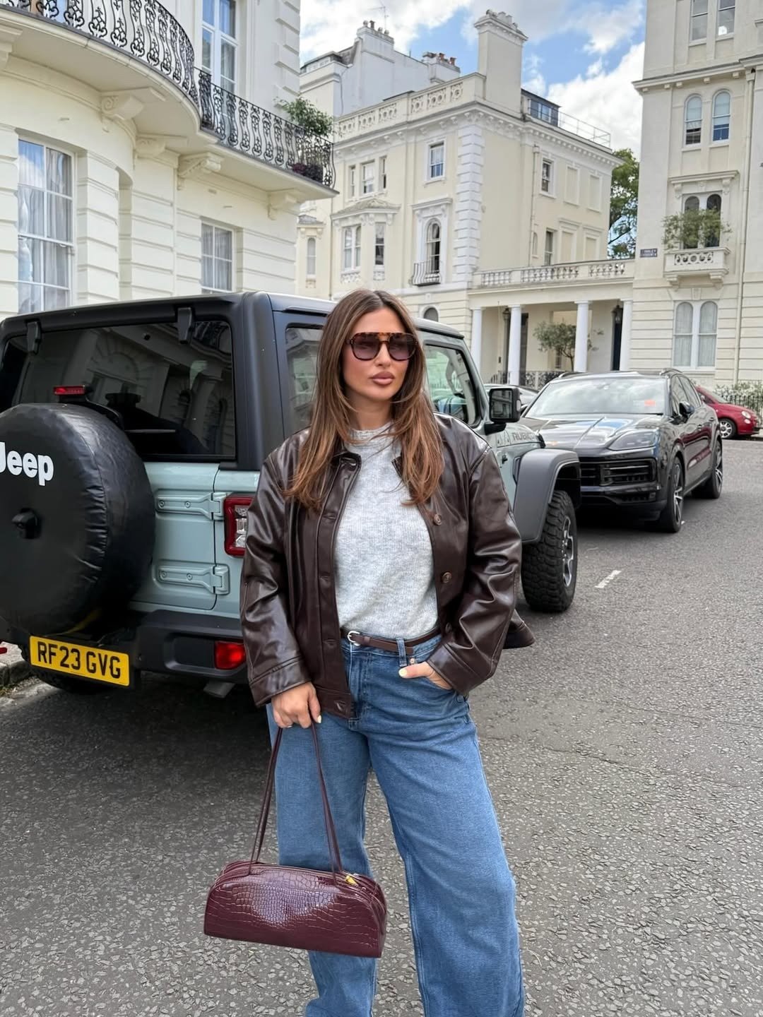 Modelo posa com jaqueta marrom, tricô cinza e jeans azul em rua elegante; sapatos confortáveis femininos não visíveis.