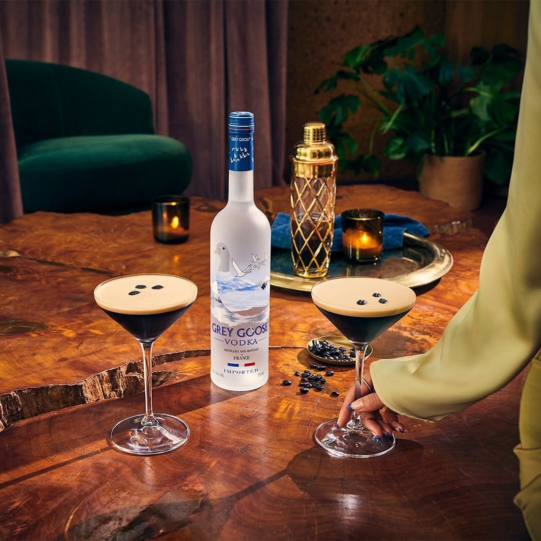 Mão com manga longa verde clara segura taça de drinks com vodka fáceis em mesa rústica, ambiente acolhedor e elegante.