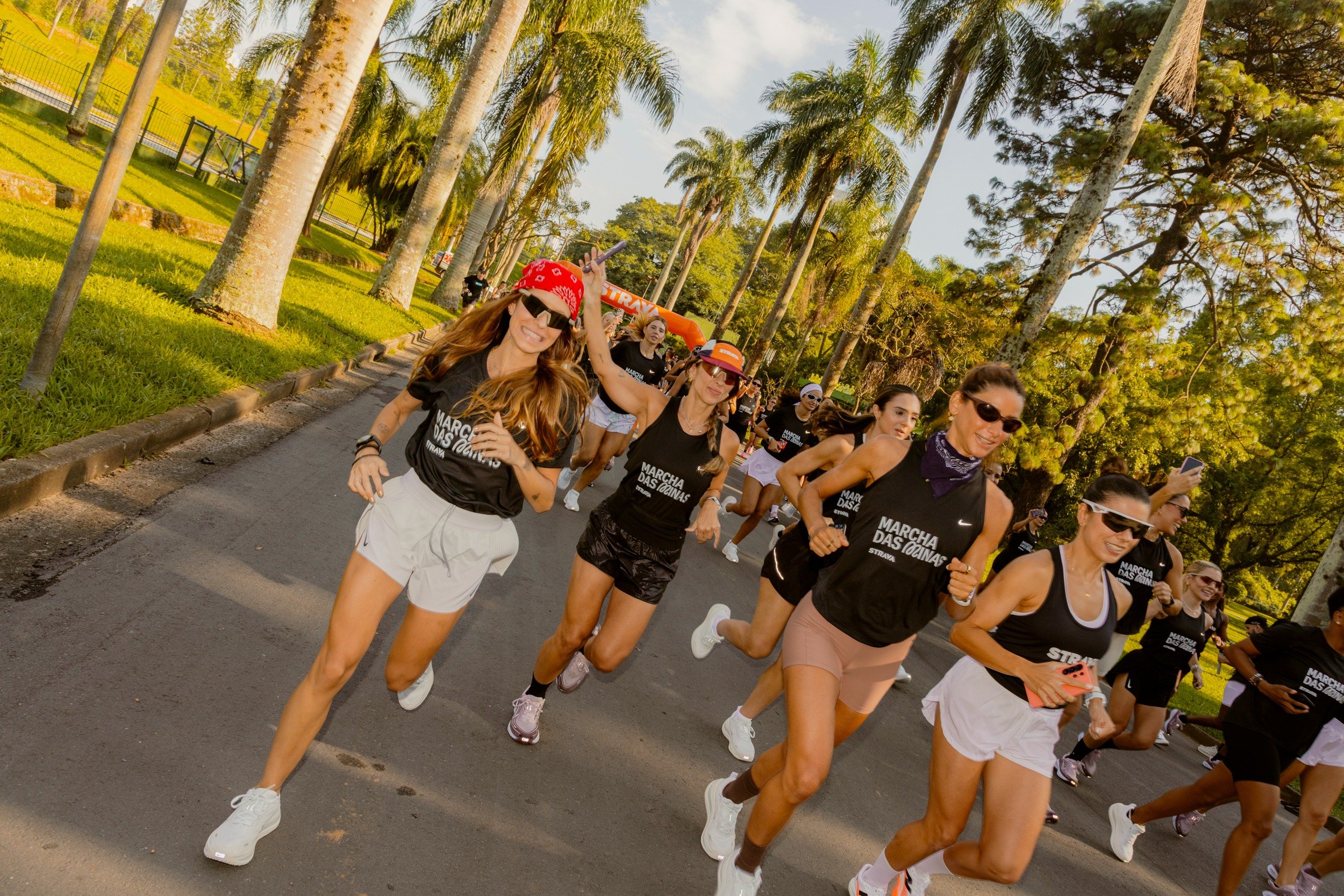 Corredoras da strava correm sorrindo com regatas pretas e shorts claros em alameda de palmeiras.